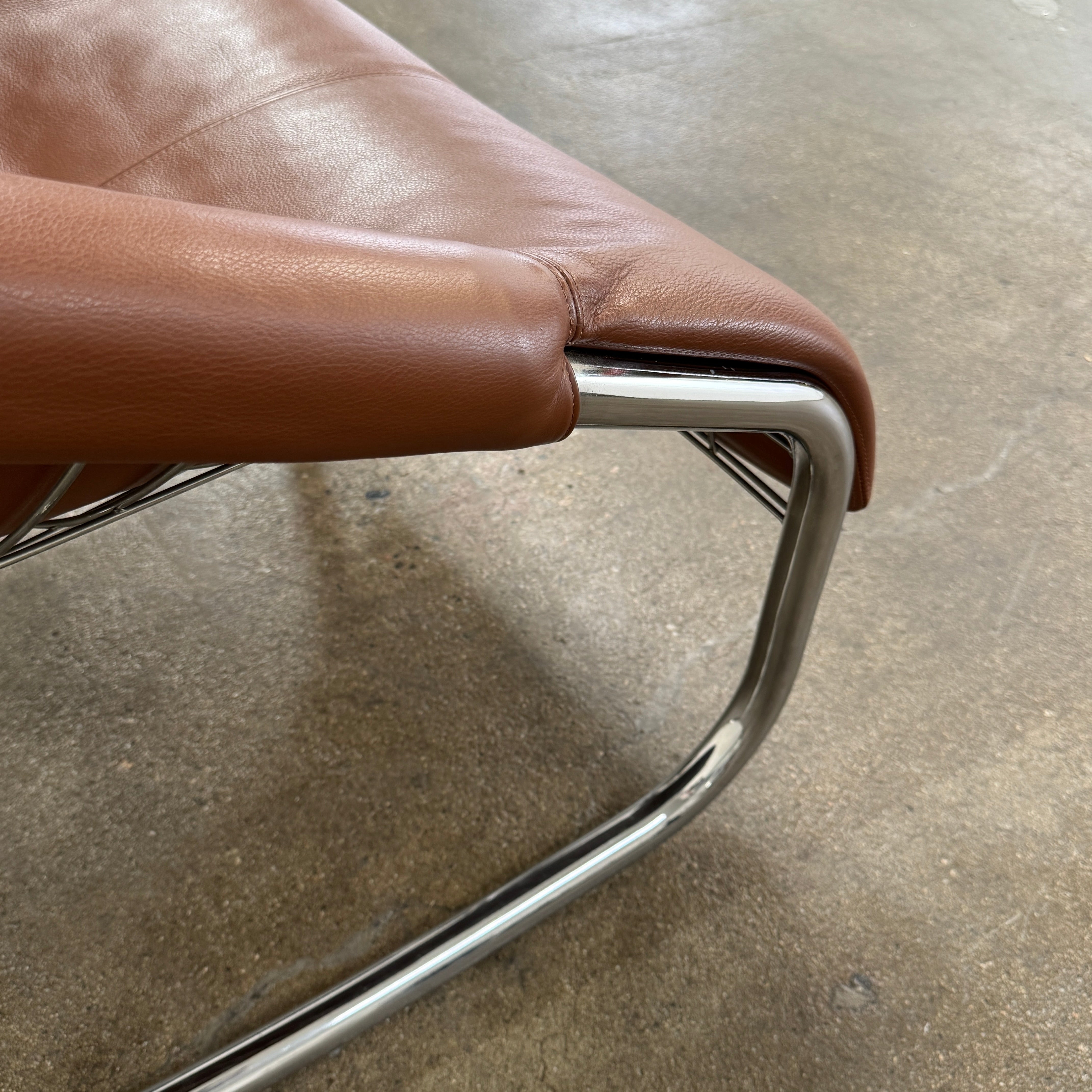 The Minotti Cortina Armchair by Minotti features luxurious brown leather upholstery and a gracefully curved seat, supported by a sleek chrome metal frame, and is showcased against a plain white background.