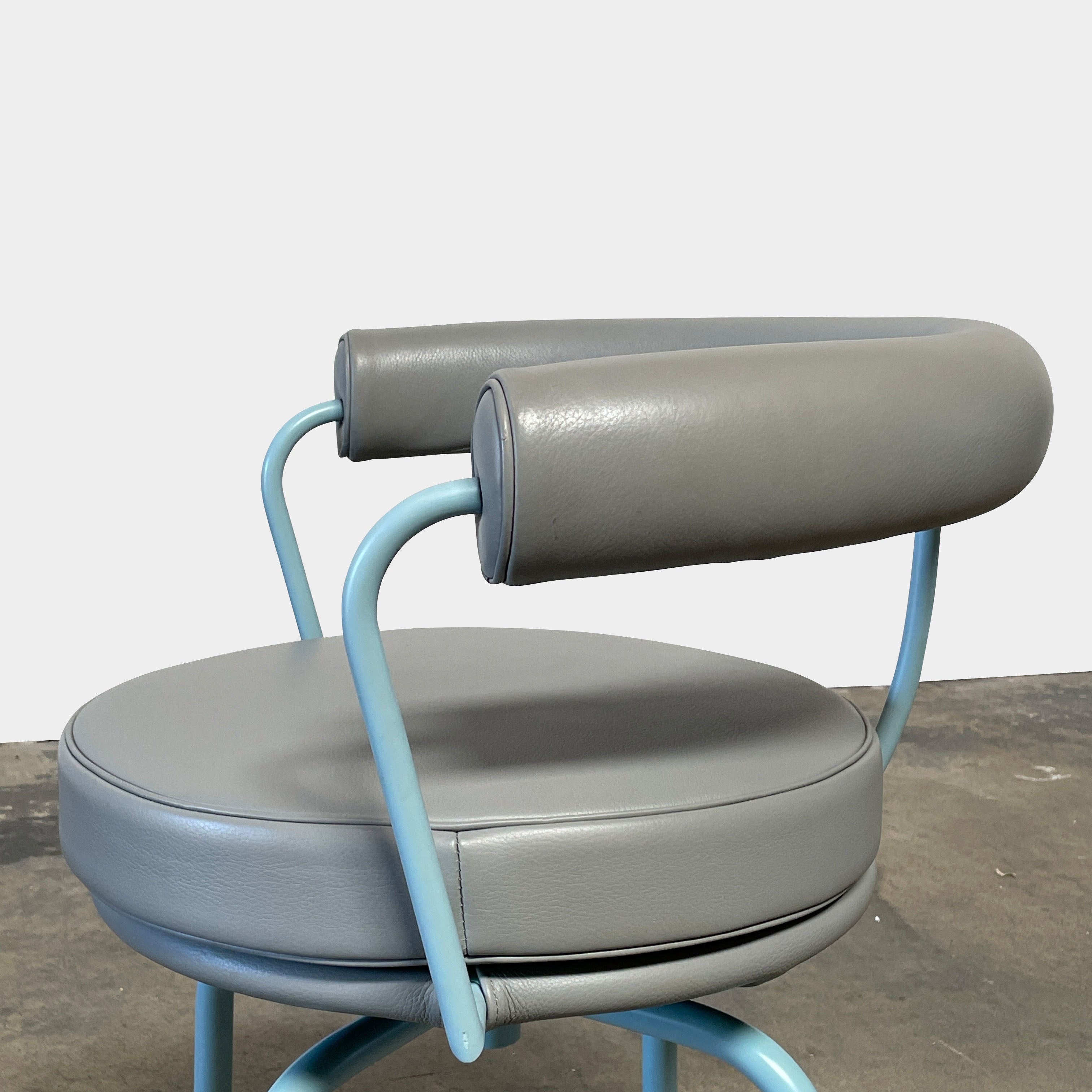 A gray leather Cassina LC7 Swivel Chair, featuring a circular seat, tubular backrest, and light blue metal legs, embodies modern design by Le Corbusier on a white background.