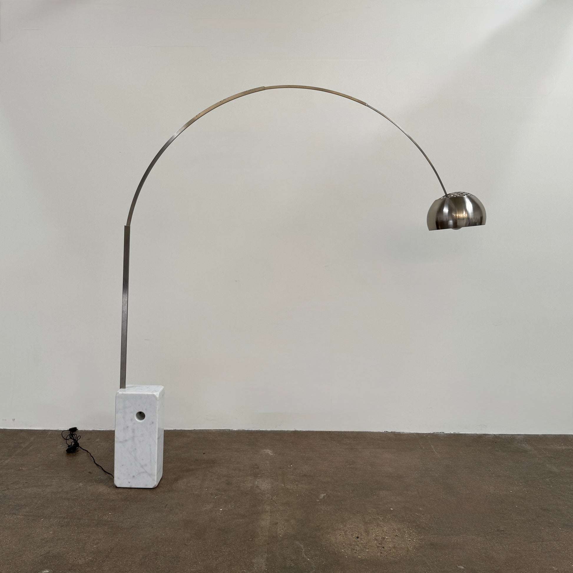 The DWR Arco Floor Lamp by DWR features a marble base, a sleek metal arching stem, and a dome-shaped metal shade; inspired by Italian craftsmanship and Achille Castiglioni’s legendary design, with an electrical cord and plug visible near the base.
