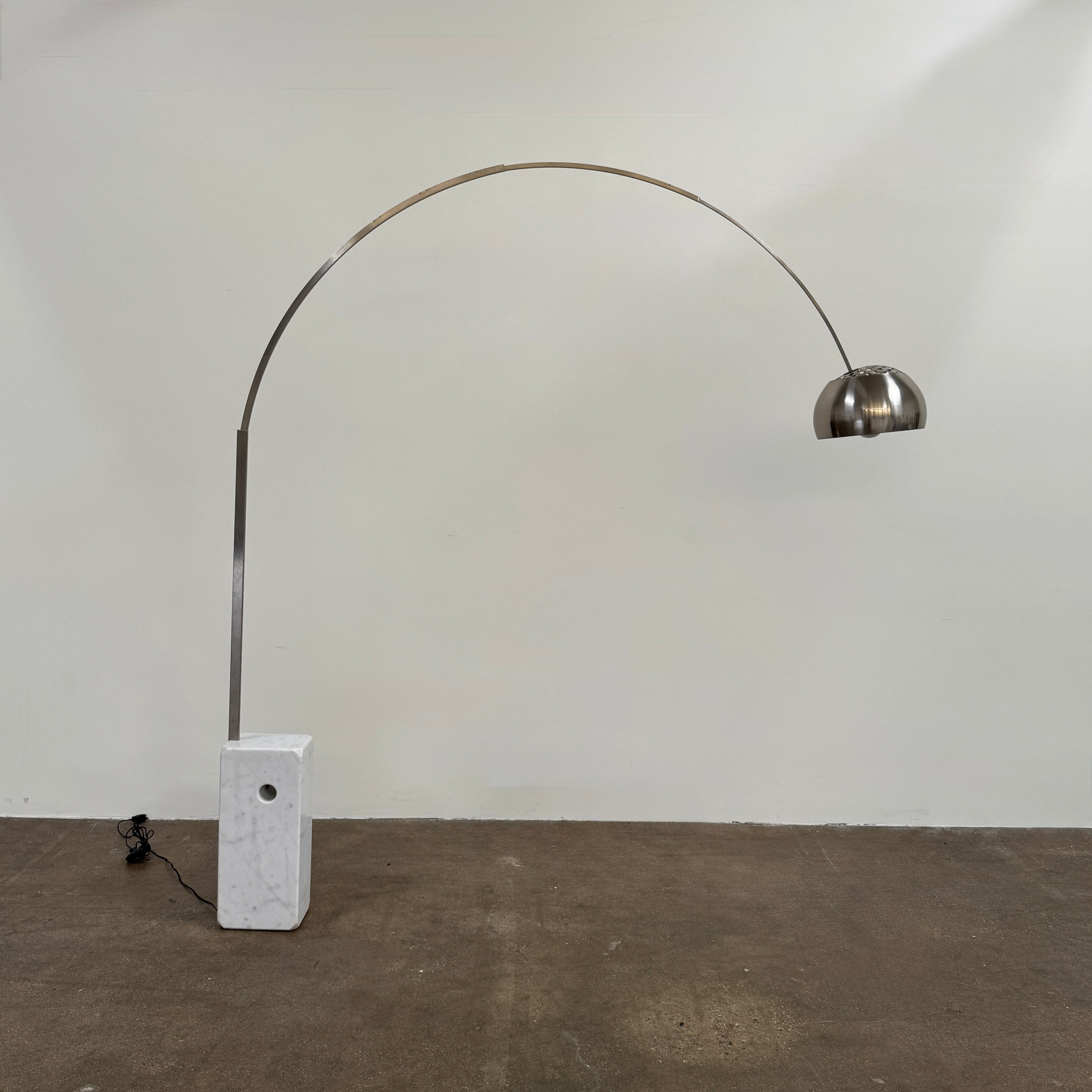 The DWR Arco Floor Lamp by DWR features a marble base, a sleek metal arching stem, and a dome-shaped metal shade; inspired by Italian craftsmanship and Achille Castiglioni’s legendary design, with an electrical cord and plug visible near the base.