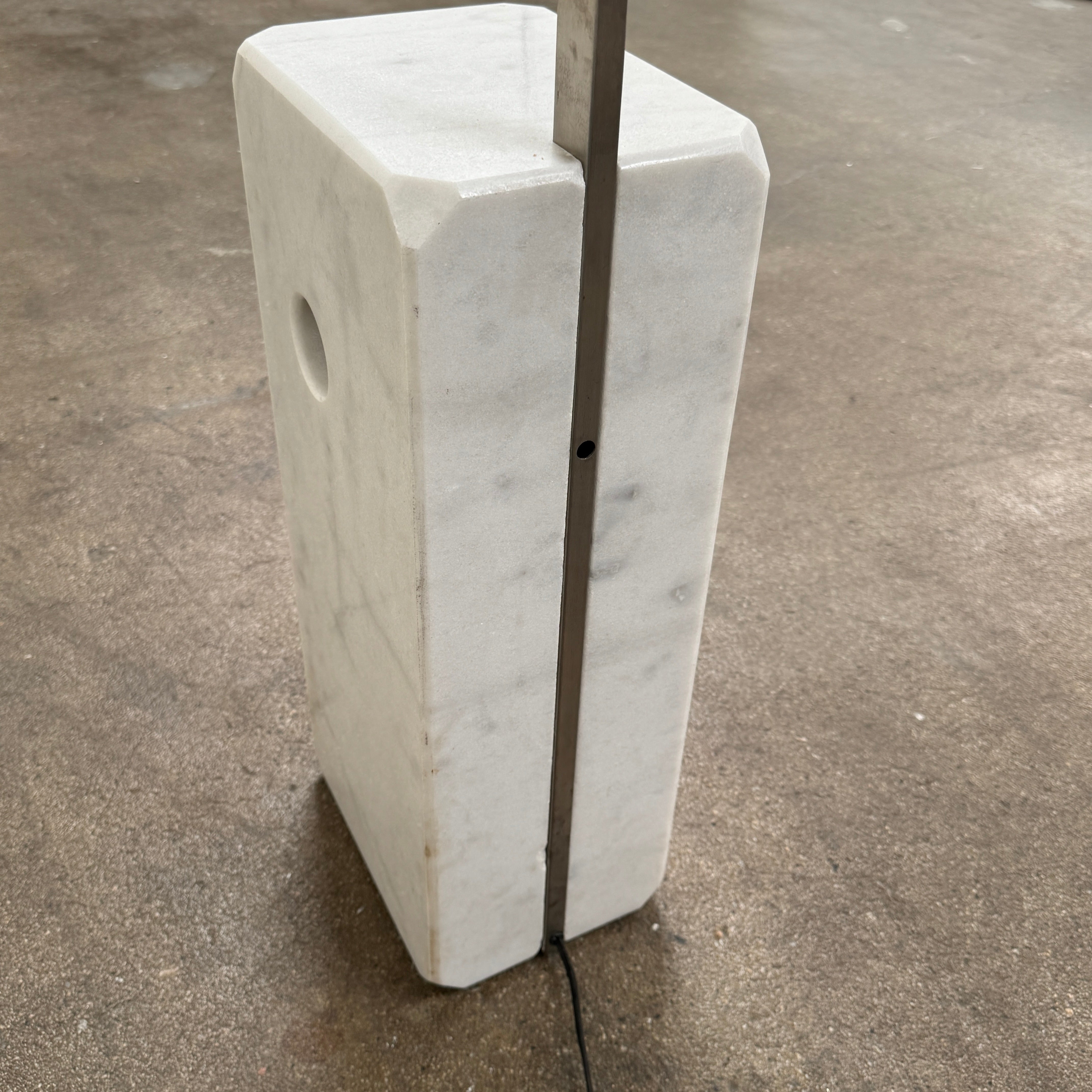 The DWR Arco Floor Lamp by DWR features a marble base, a sleek metal arching stem, and a dome-shaped metal shade; inspired by Italian craftsmanship and Achille Castiglioni’s legendary design, with an electrical cord and plug visible near the base.