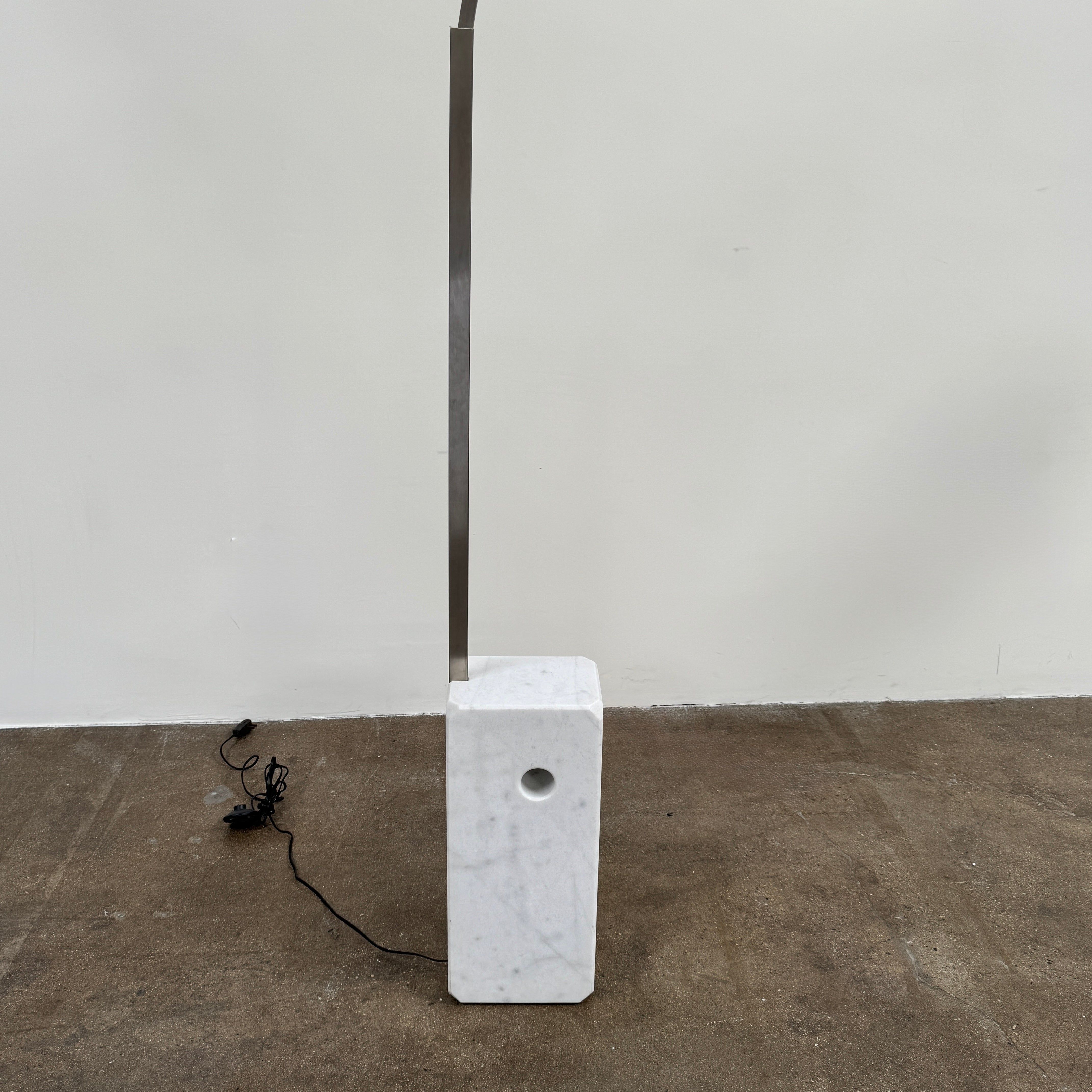 The DWR Arco Floor Lamp by DWR features a marble base, a sleek metal arching stem, and a dome-shaped metal shade; inspired by Italian craftsmanship and Achille Castiglioni’s legendary design, with an electrical cord and plug visible near the base.