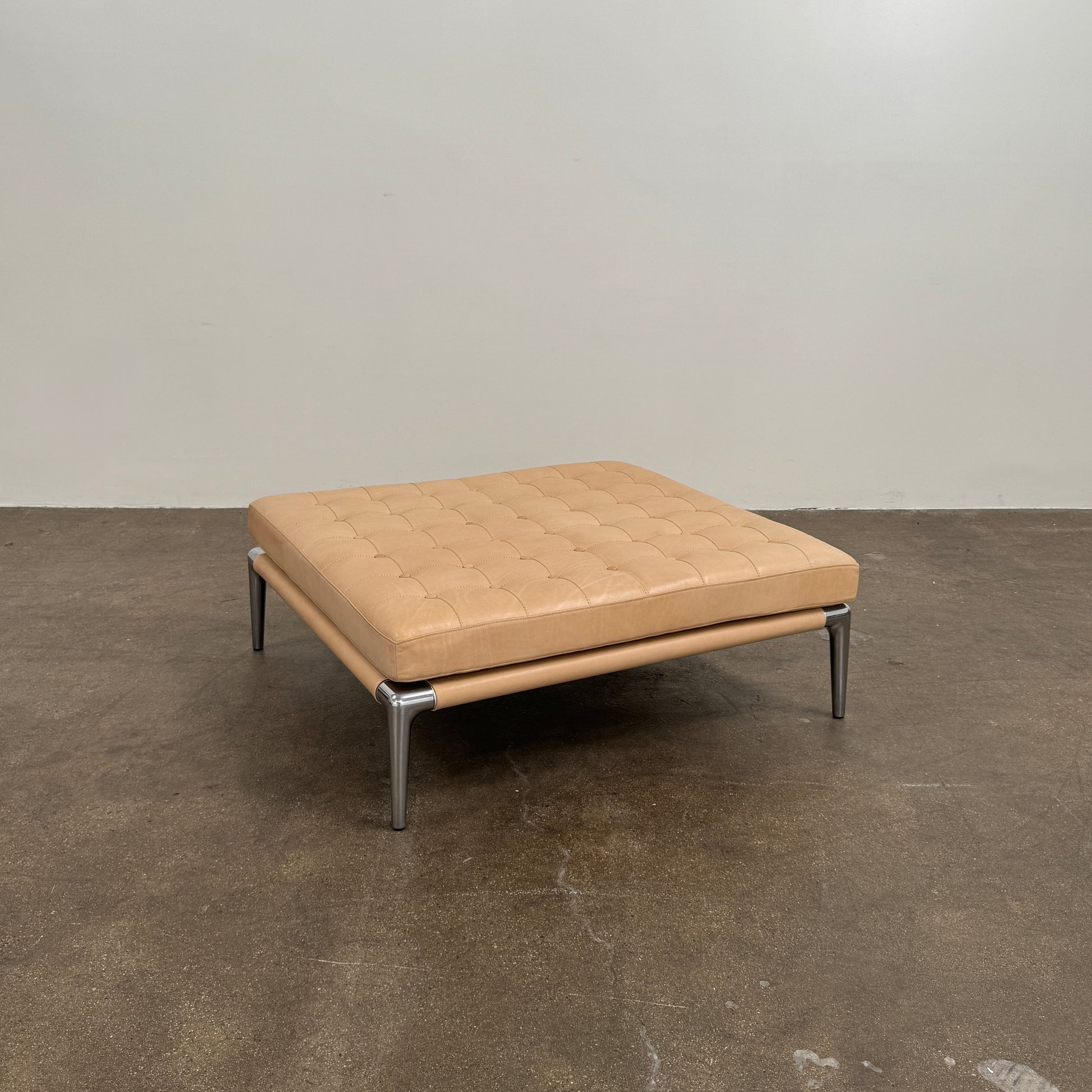 The Cassina Prive Ottoman by Cassina is a rectangular tan leather luxury ottoman featuring tufted upholstery and sleek metal legs, displayed against a plain white background.