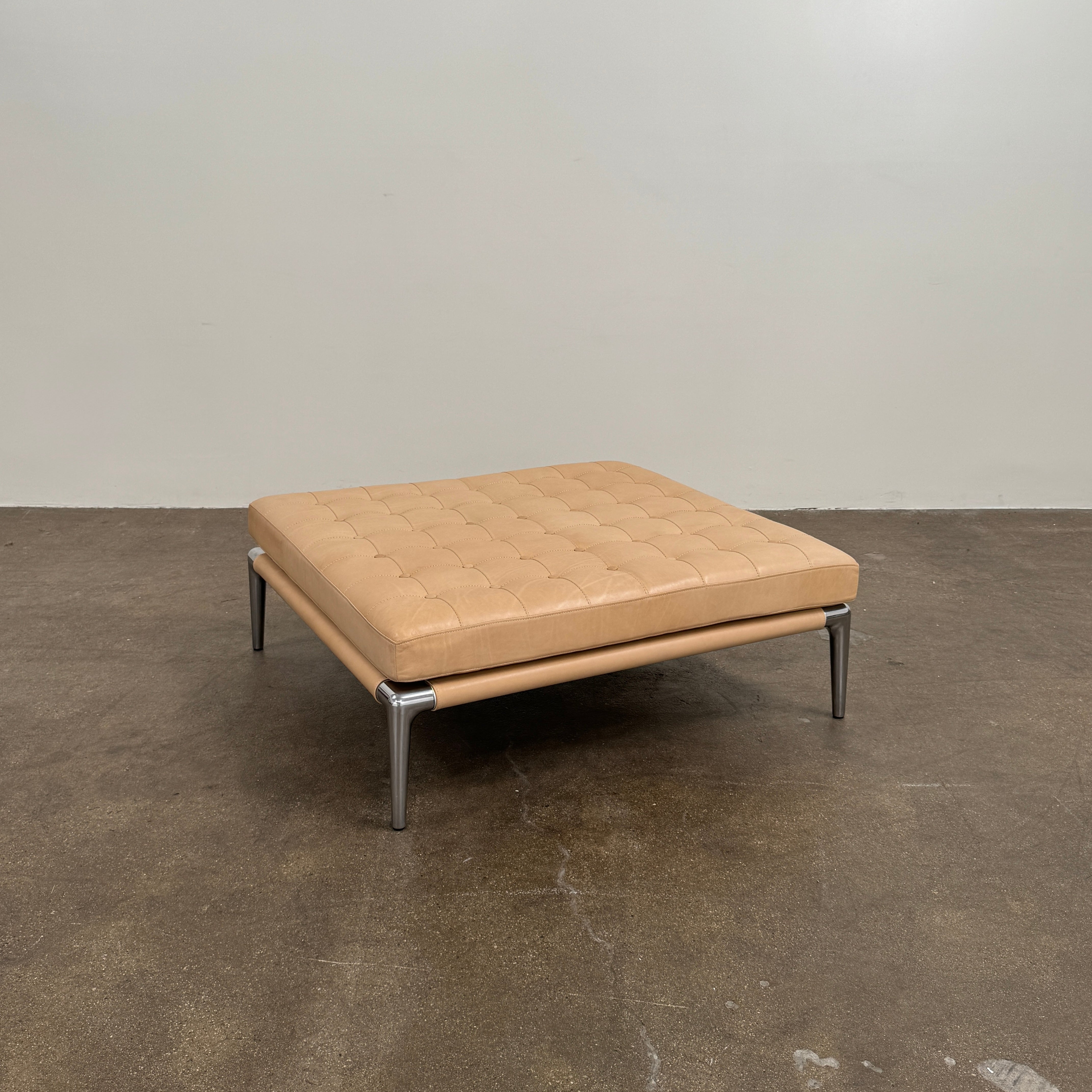 The Cassina Prive Ottoman by Cassina is a rectangular tan leather luxury ottoman featuring tufted upholstery and sleek metal legs, displayed against a plain white background.