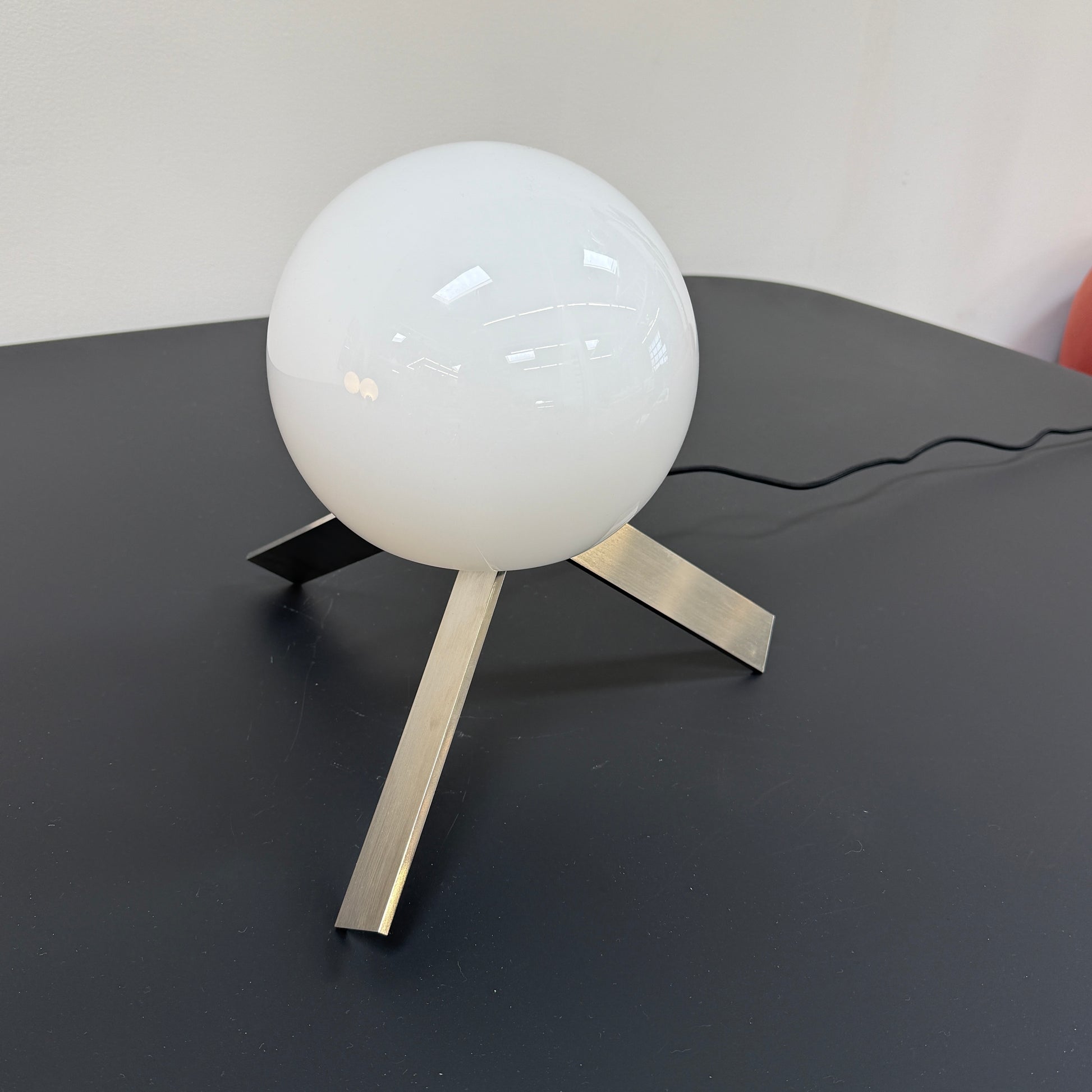 The Michael Anastassiades To The Top Table Lamp features a spherical white opaline glass shade and sits on three gold angled legs, designed by Michael Anastassiades, set against a plain light background.