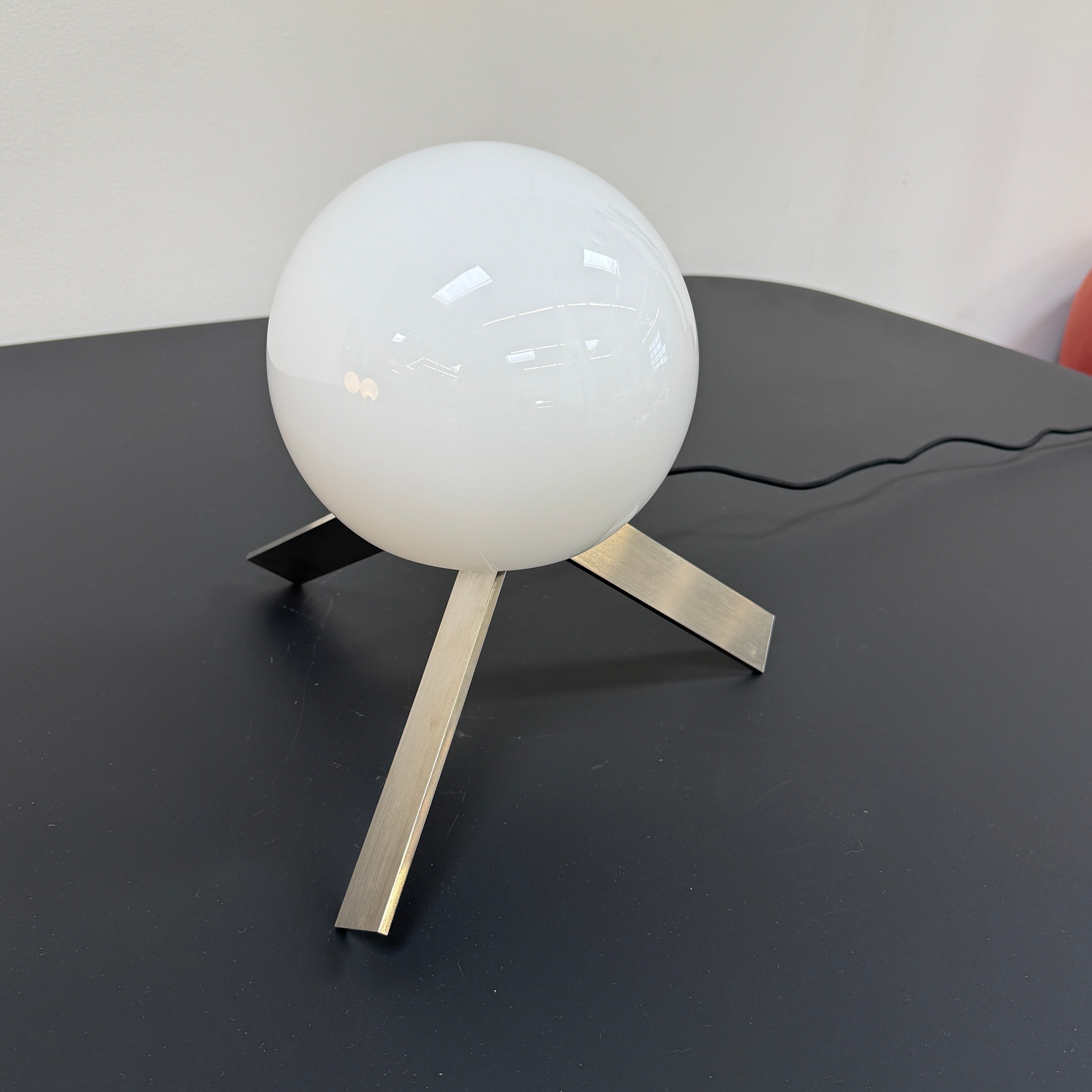 The Michael Anastassiades To The Top Table Lamp features a spherical white opaline glass shade and sits on three gold angled legs, designed by Michael Anastassiades, set against a plain light background.