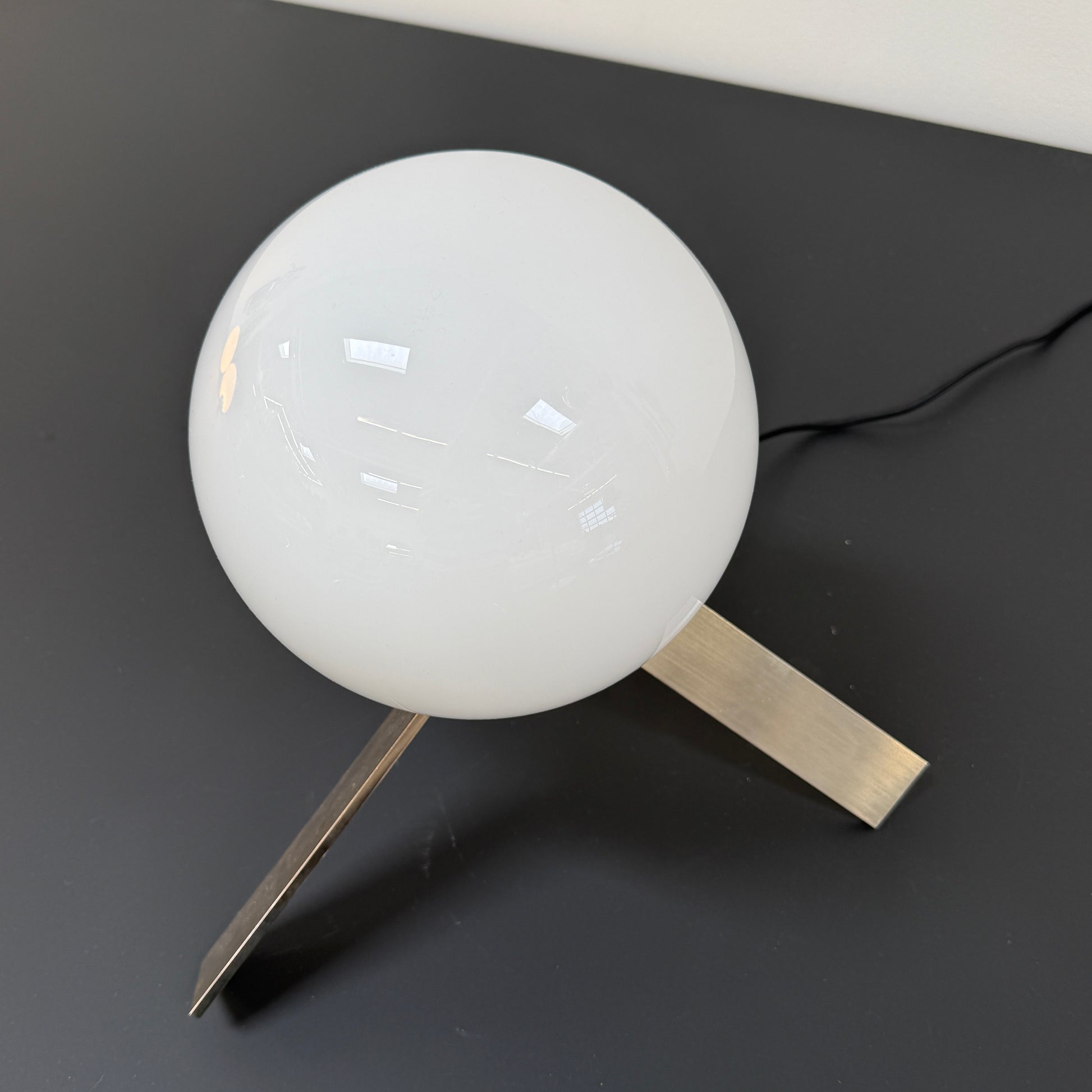 The Michael Anastassiades To The Top Table Lamp features a spherical white opaline glass shade and sits on three gold angled legs, designed by Michael Anastassiades, set against a plain light background.