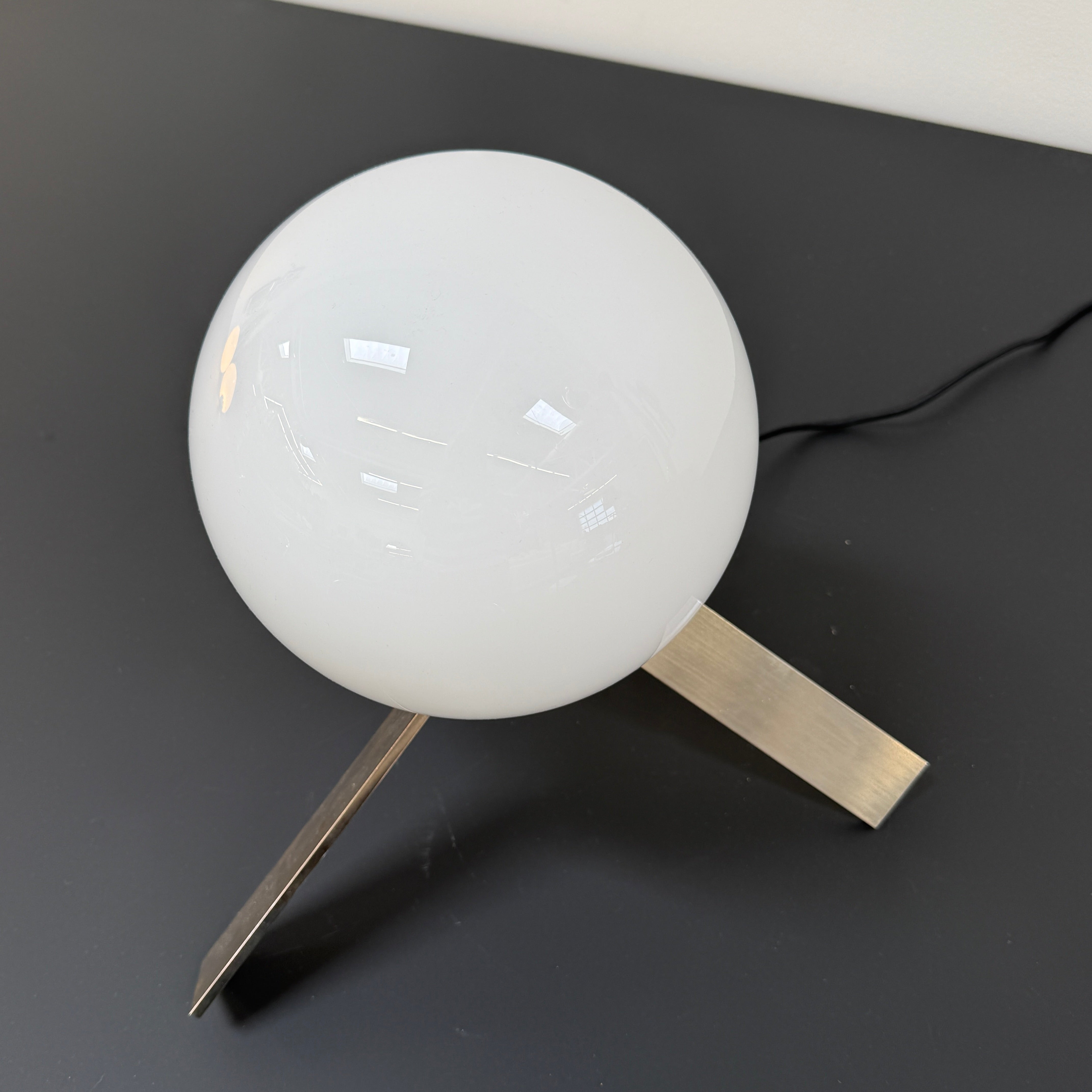 The Michael Anastassiades To The Top Table Lamp features a spherical white opaline glass shade and sits on three gold angled legs, designed by Michael Anastassiades, set against a plain light background.