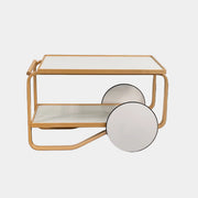 The Artek Aalto Tea Trolley 901 by Artek is a wooden bar cart inspired by the iconic Aalto Tea Trolley, showcasing two large white wheels, a handle, and two white shelves. Set against a plain white background, it stands as a testament to handcrafted Finnish design in the spirit of Alvar Aalto.