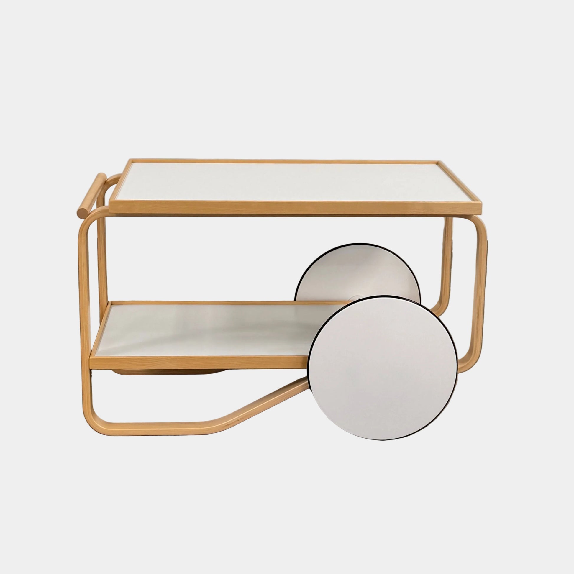 The Artek Aalto Tea Trolley 901 by Artek is a wooden bar cart inspired by the iconic Aalto Tea Trolley, showcasing two large white wheels, a handle, and two white shelves. Set against a plain white background, it stands as a testament to handcrafted Finnish design in the spirit of Alvar Aalto.