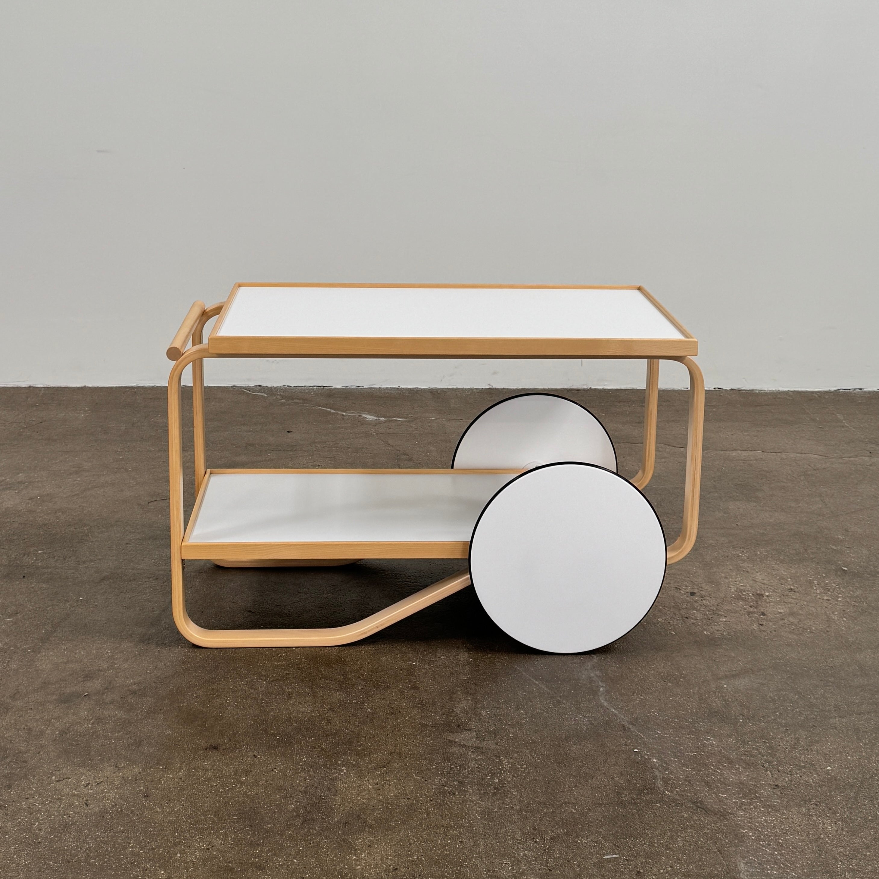 The Artek Aalto Tea Trolley 901 by Artek is a wooden bar cart inspired by the iconic Aalto Tea Trolley, showcasing two large white wheels, a handle, and two white shelves. Set against a plain white background, it stands as a testament to handcrafted Finnish design in the spirit of Alvar Aalto.