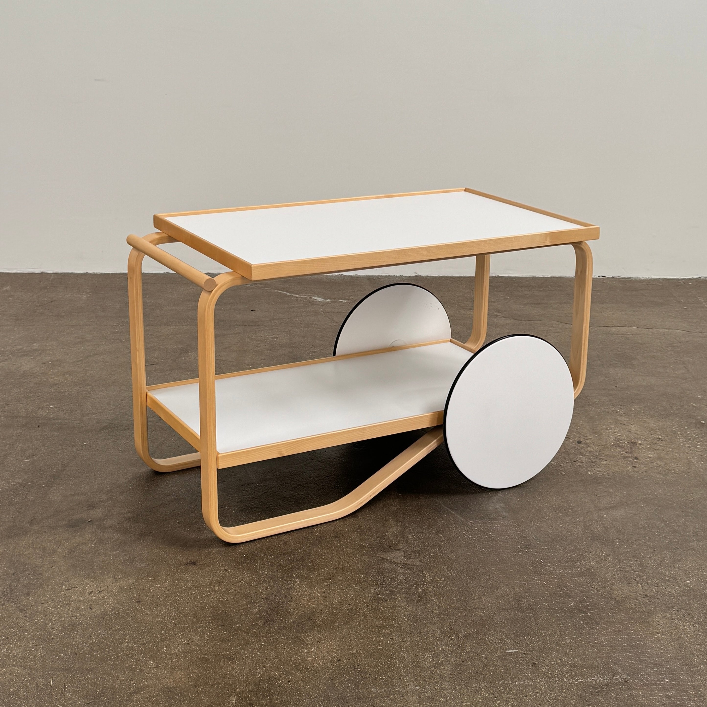 The Artek Aalto Tea Trolley 901 by Artek is a wooden bar cart inspired by the iconic Aalto Tea Trolley, showcasing two large white wheels, a handle, and two white shelves. Set against a plain white background, it stands as a testament to handcrafted Finnish design in the spirit of Alvar Aalto.