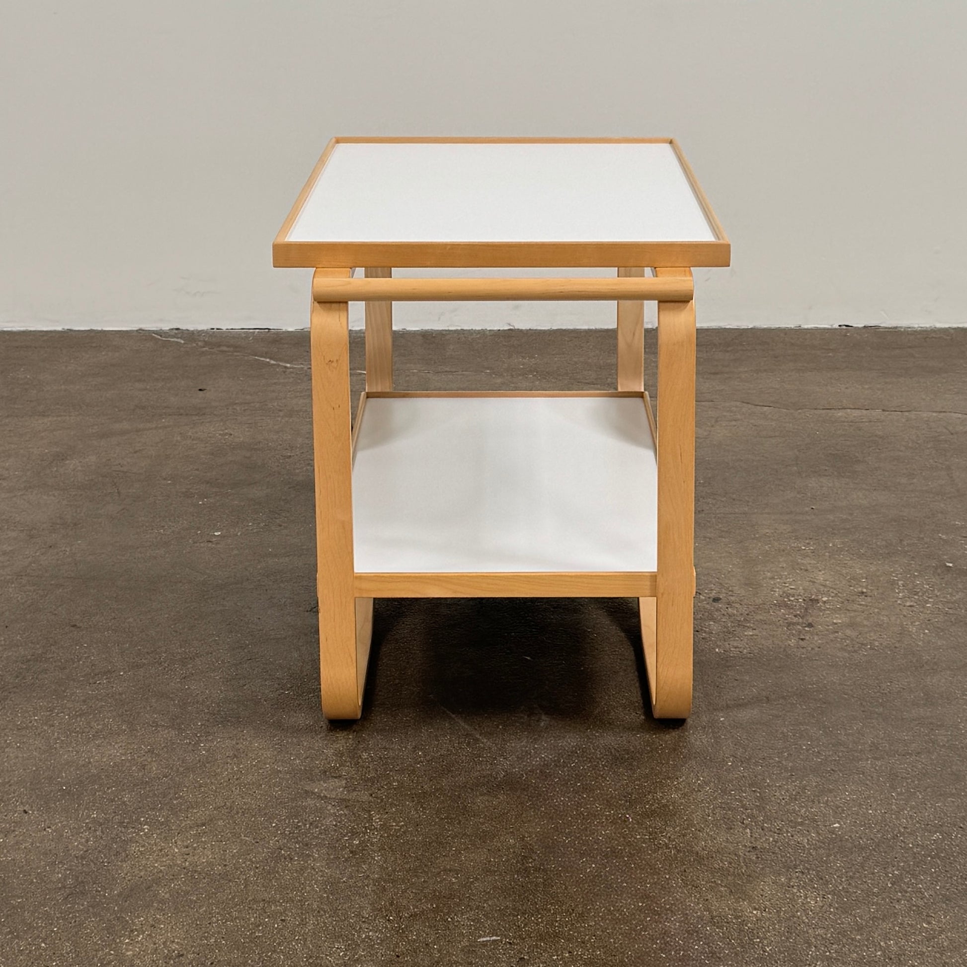 The Artek Aalto Tea Trolley 901 by Artek is a wooden bar cart inspired by the iconic Aalto Tea Trolley, showcasing two large white wheels, a handle, and two white shelves. Set against a plain white background, it stands as a testament to handcrafted Finnish design in the spirit of Alvar Aalto.