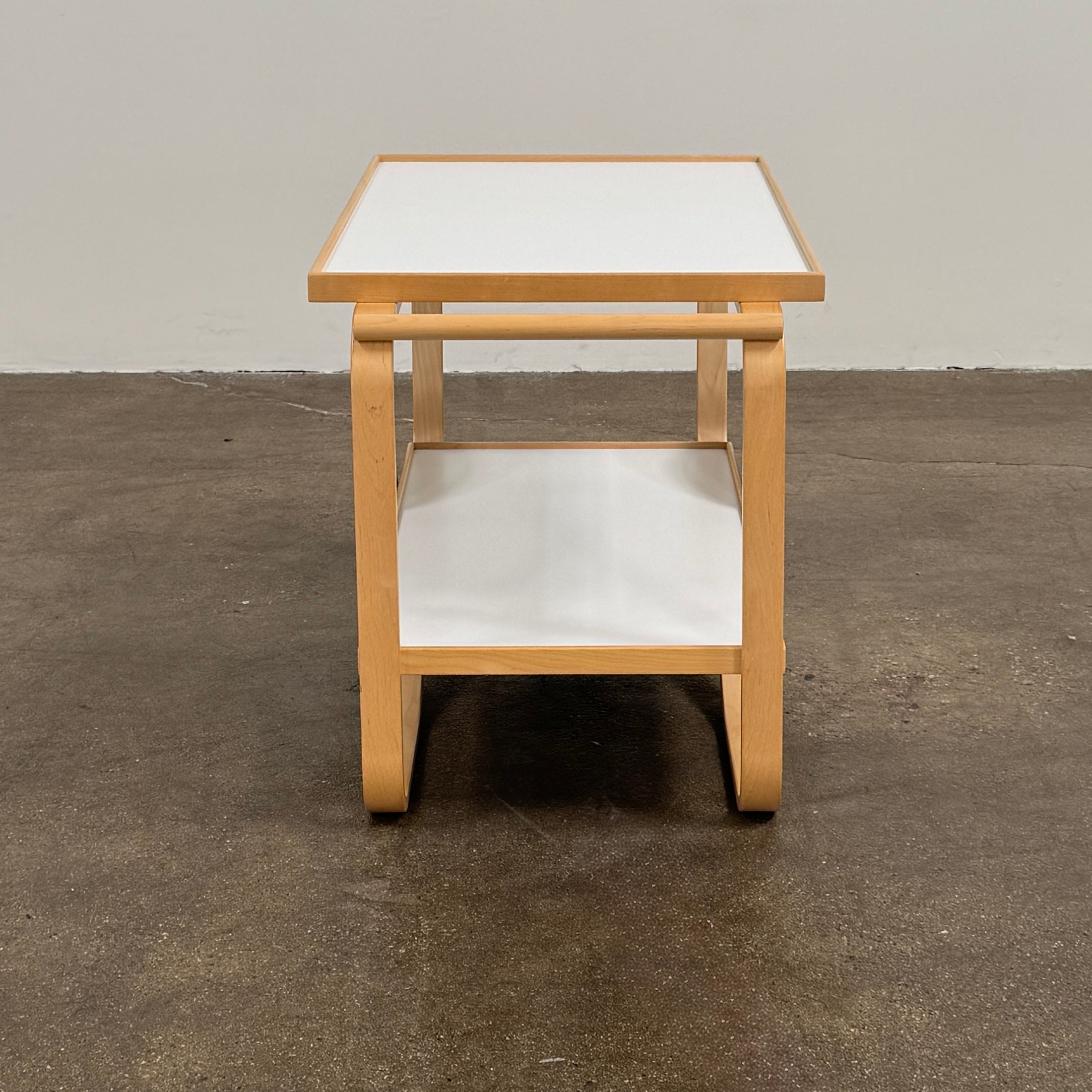The Artek Aalto Tea Trolley 901 by Artek is a wooden bar cart inspired by the iconic Aalto Tea Trolley, showcasing two large white wheels, a handle, and two white shelves. Set against a plain white background, it stands as a testament to handcrafted Finnish design in the spirit of Alvar Aalto.