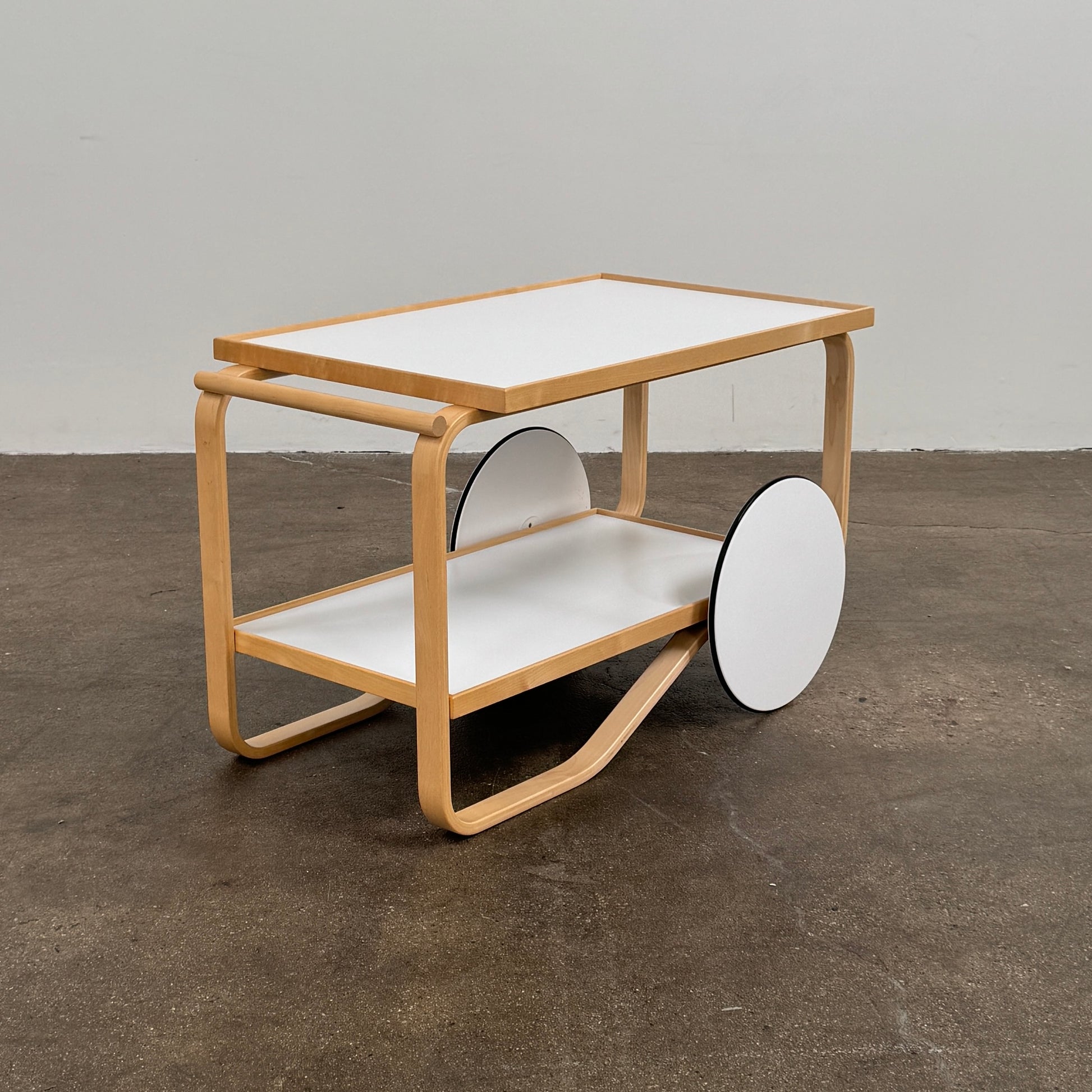 The Artek Aalto Tea Trolley 901 by Artek is a wooden bar cart inspired by the iconic Aalto Tea Trolley, showcasing two large white wheels, a handle, and two white shelves. Set against a plain white background, it stands as a testament to handcrafted Finnish design in the spirit of Alvar Aalto.