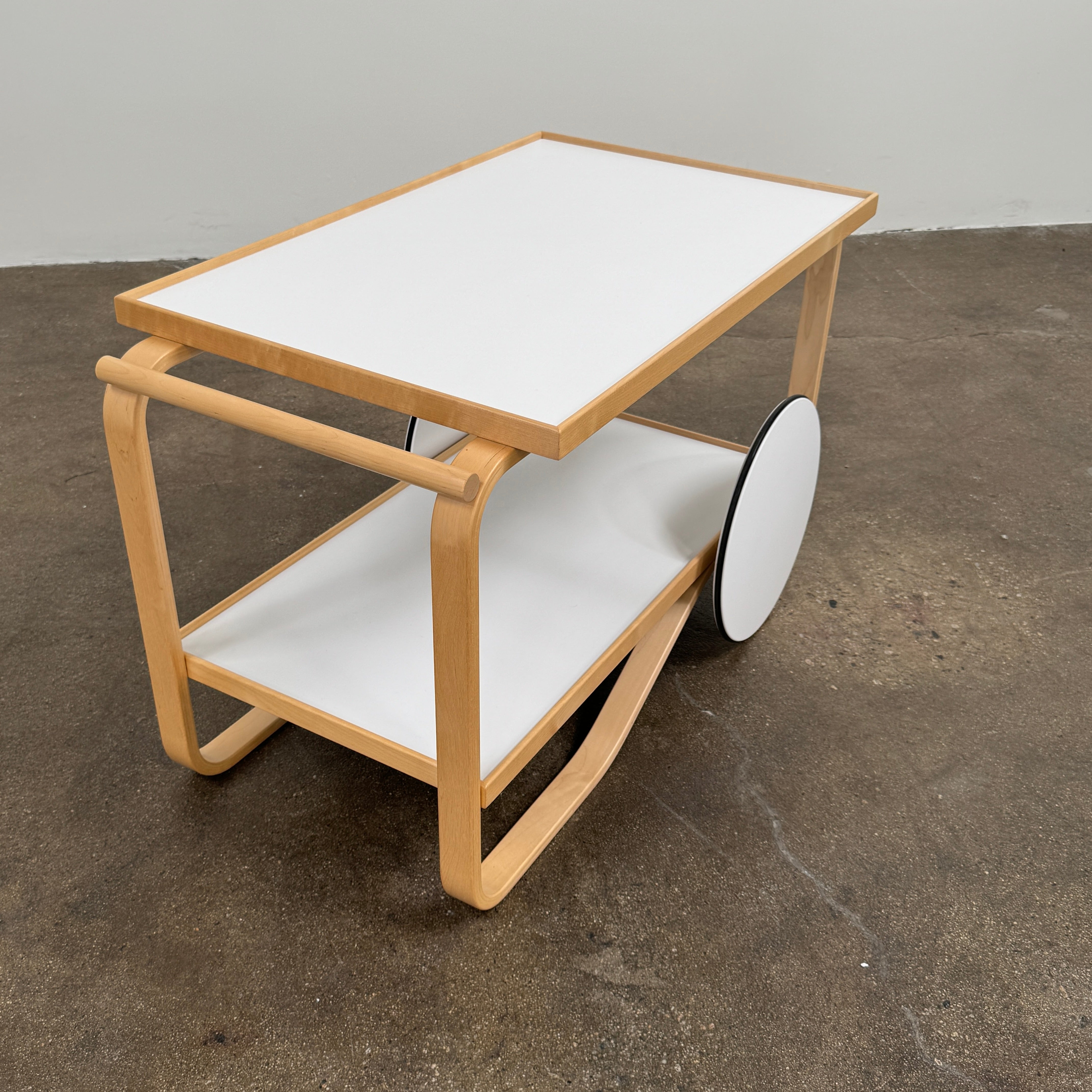 The Artek Aalto Tea Trolley 901 by Artek is a wooden bar cart inspired by the iconic Aalto Tea Trolley, showcasing two large white wheels, a handle, and two white shelves. Set against a plain white background, it stands as a testament to handcrafted Finnish design in the spirit of Alvar Aalto.