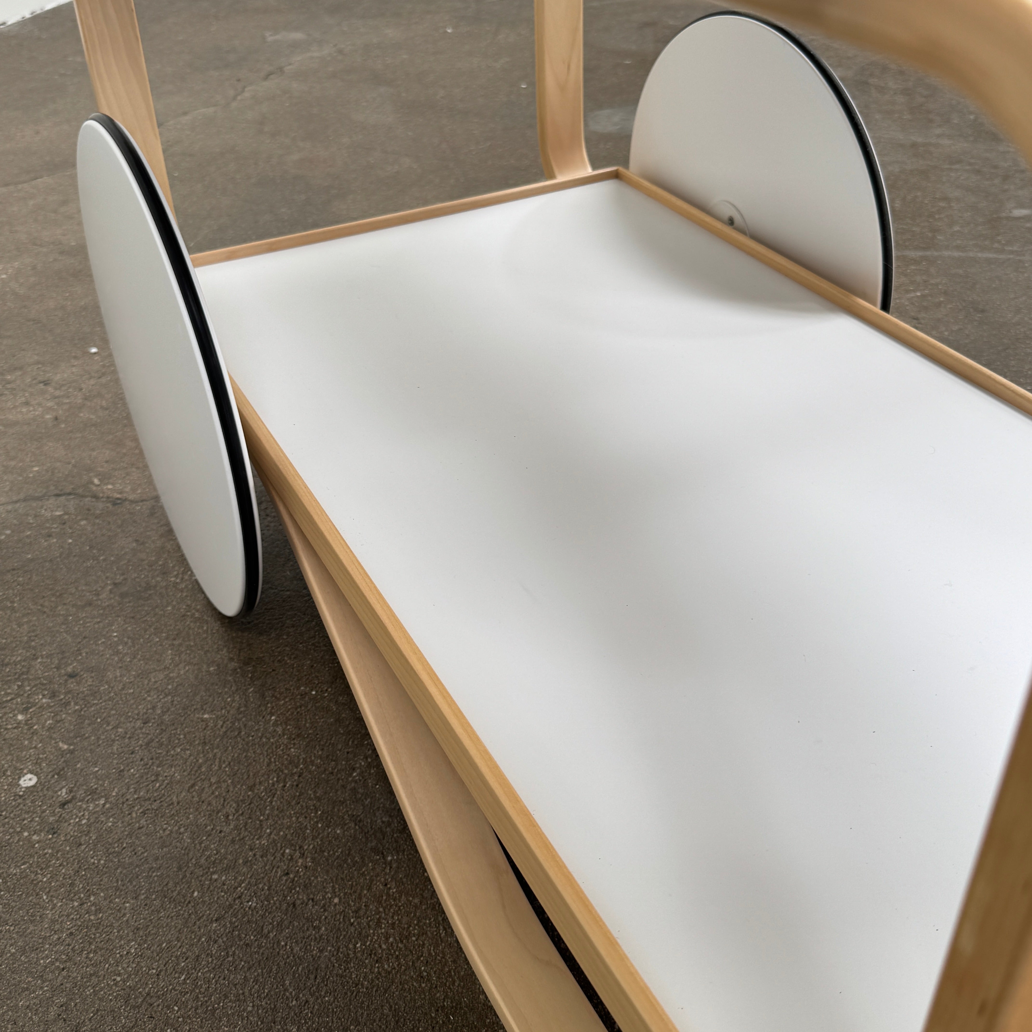 The Artek Aalto Tea Trolley 901 by Artek is a wooden bar cart inspired by the iconic Aalto Tea Trolley, showcasing two large white wheels, a handle, and two white shelves. Set against a plain white background, it stands as a testament to handcrafted Finnish design in the spirit of Alvar Aalto.