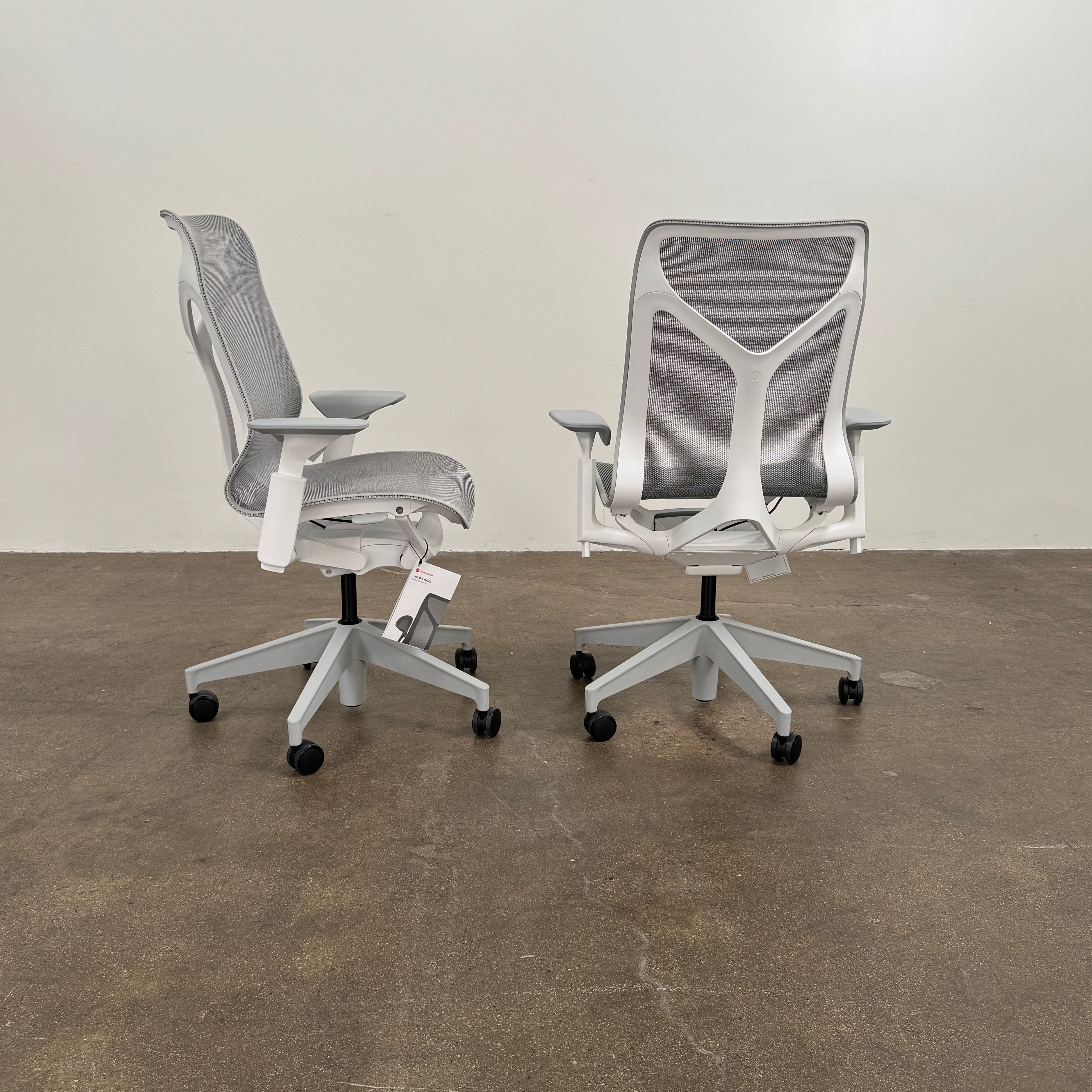 A sleek, modern white Herman Miller Cosm Office Chair featuring a mesh back and seat, supportive armrests, and five caster wheels is showcased on a concrete floor against an off-white wall. Designed by Herman Miller, this ergonomic chair seamlessly combines contemporary style with exceptional comfort.