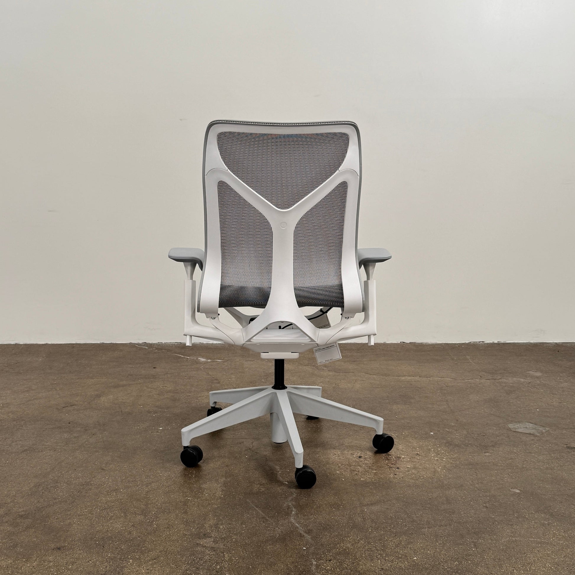 A sleek, modern white Herman Miller Cosm Office Chair featuring a mesh back and seat, supportive armrests, and five caster wheels is showcased on a concrete floor against an off-white wall. Designed by Herman Miller, this ergonomic chair seamlessly combines contemporary style with exceptional comfort.