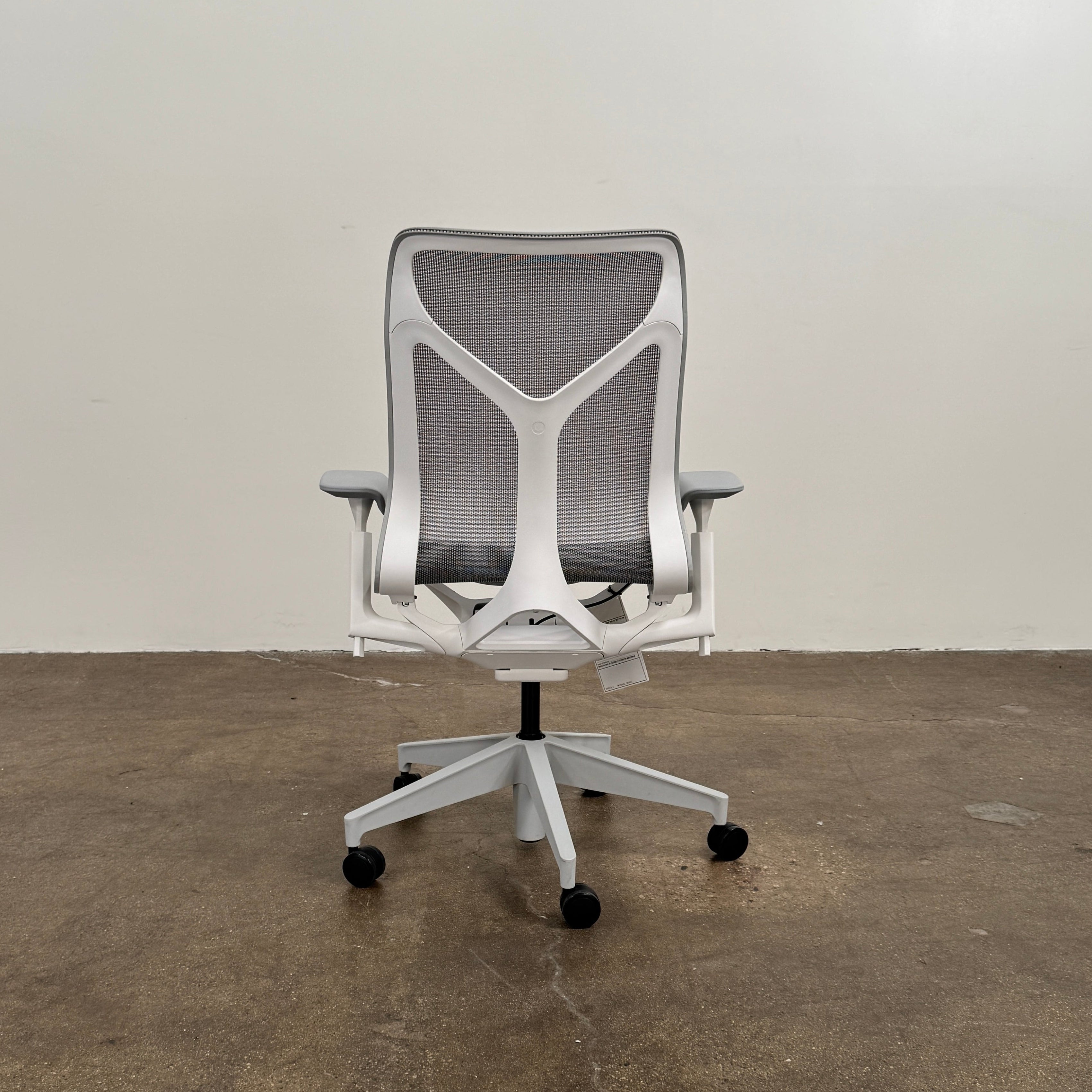 A sleek, modern white Herman Miller Cosm Office Chair featuring a mesh back and seat, supportive armrests, and five caster wheels is showcased on a concrete floor against an off-white wall. Designed by Herman Miller, this ergonomic chair seamlessly combines contemporary style with exceptional comfort.