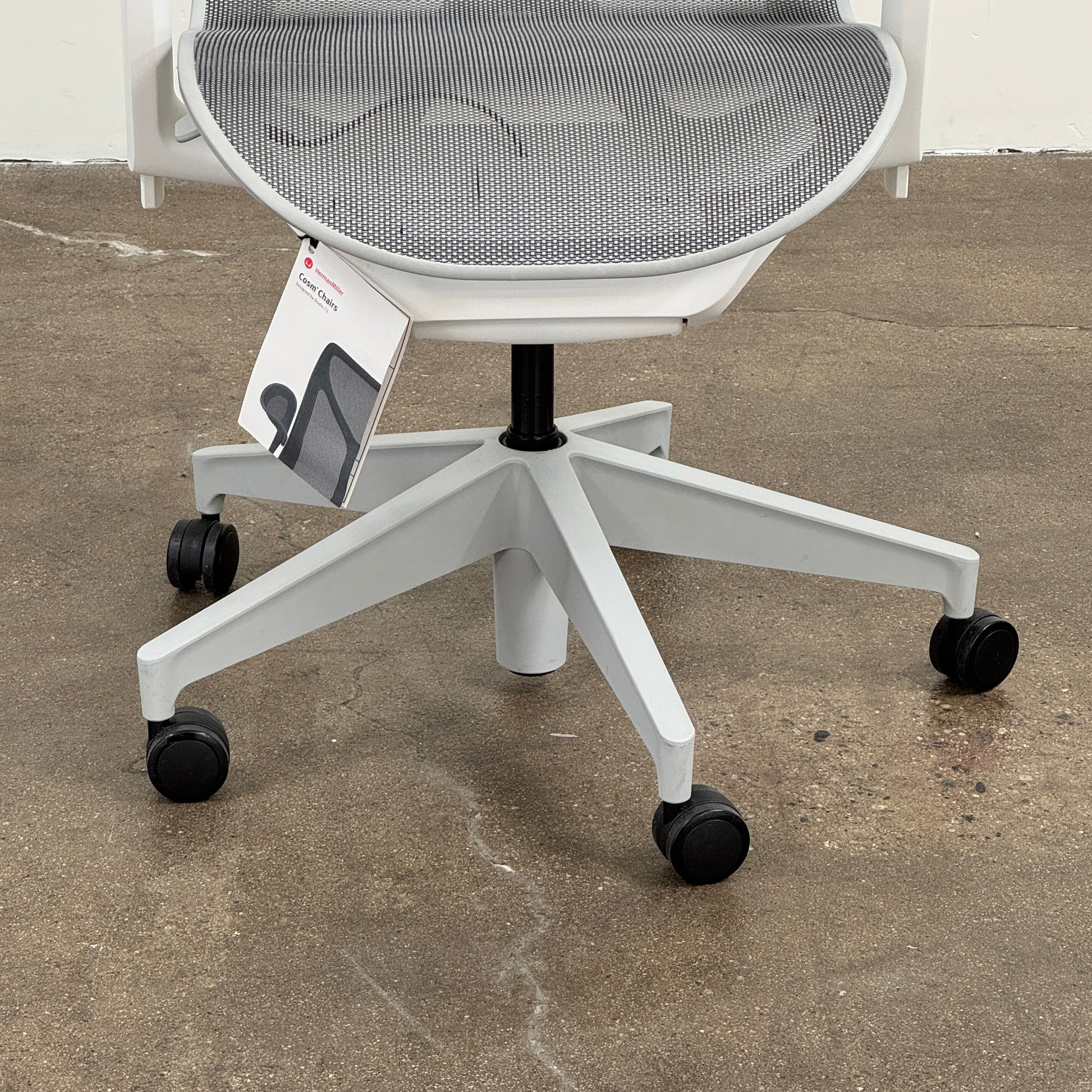 A sleek, modern white Herman Miller Cosm Office Chair featuring a mesh back and seat, supportive armrests, and five caster wheels is showcased on a concrete floor against an off-white wall. Designed by Herman Miller, this ergonomic chair seamlessly combines contemporary style with exceptional comfort.