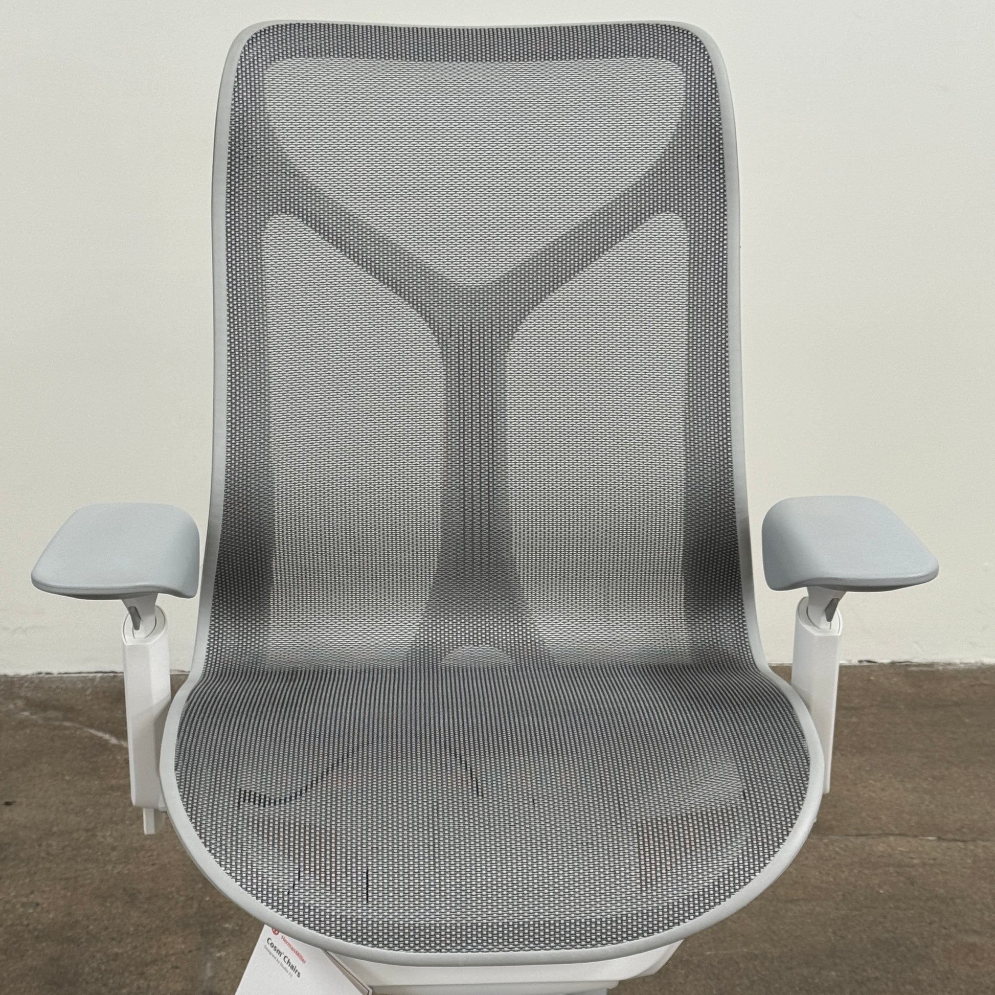 A sleek, modern white Herman Miller Cosm Office Chair featuring a mesh back and seat, supportive armrests, and five caster wheels is showcased on a concrete floor against an off-white wall. Designed by Herman Miller, this ergonomic chair seamlessly combines contemporary style with exceptional comfort.
