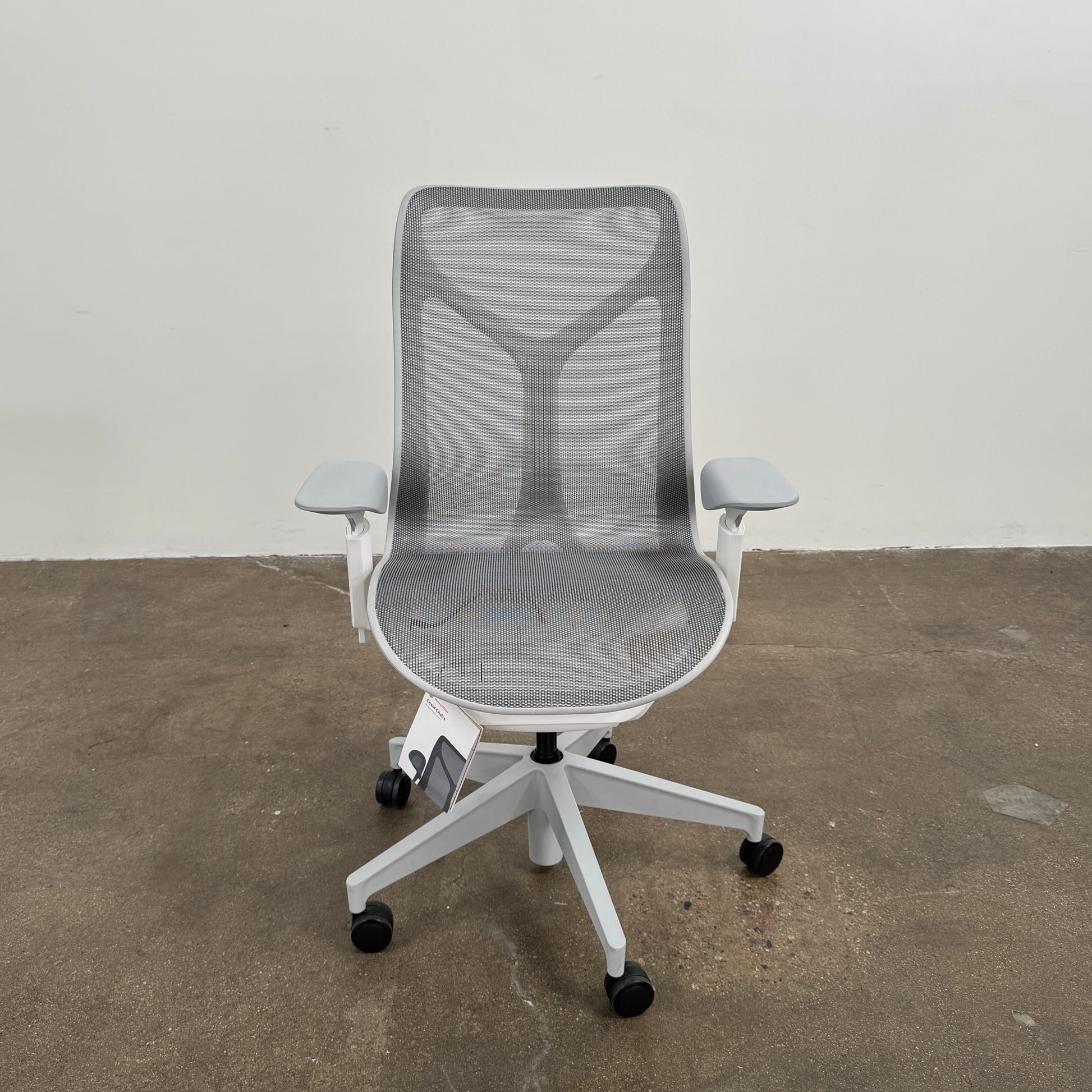 A sleek, modern white Herman Miller Cosm Office Chair featuring a mesh back and seat, supportive armrests, and five caster wheels is showcased on a concrete floor against an off-white wall. Designed by Herman Miller, this ergonomic chair seamlessly combines contemporary style with exceptional comfort.