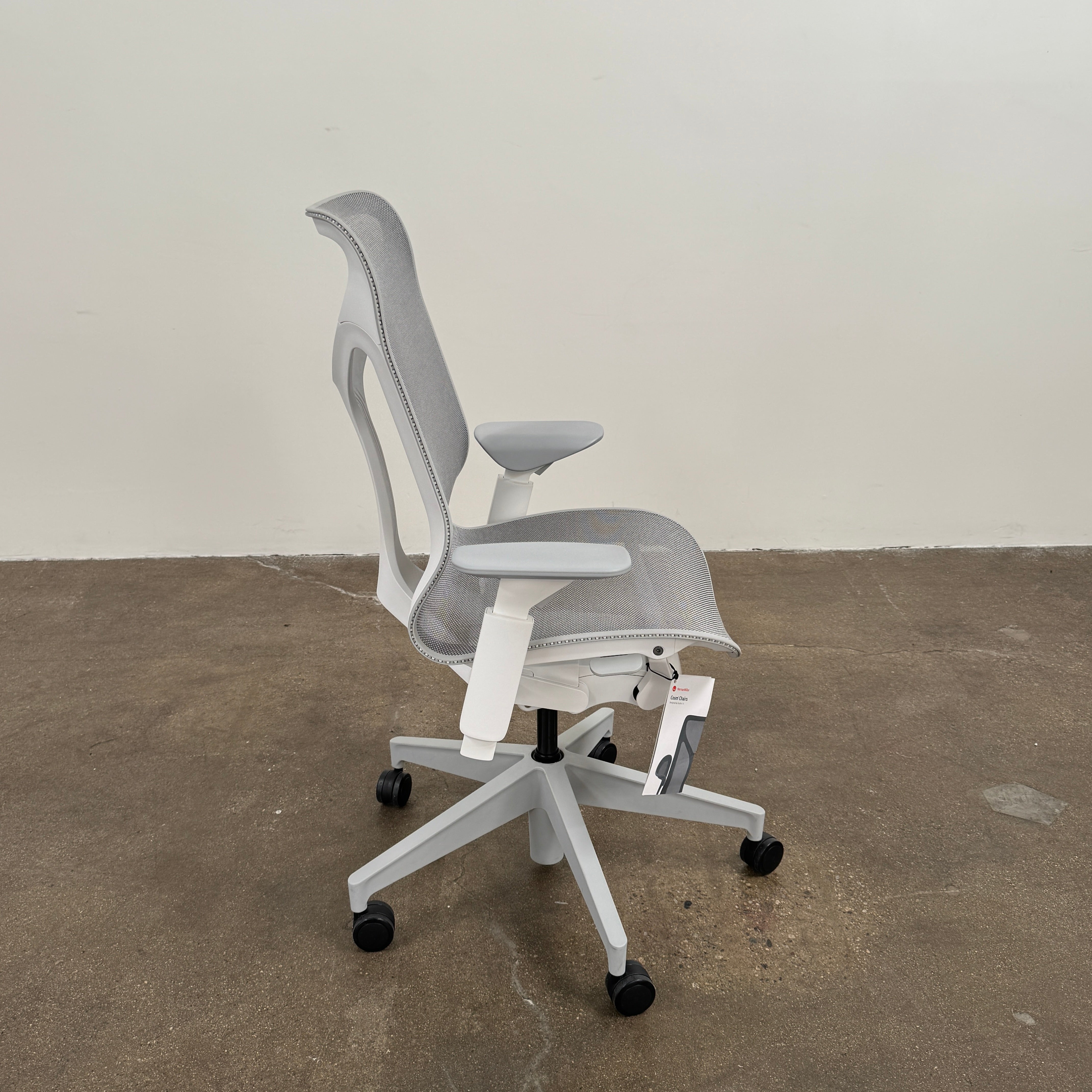 A sleek, modern white Herman Miller Cosm Office Chair featuring a mesh back and seat, supportive armrests, and five caster wheels is showcased on a concrete floor against an off-white wall. Designed by Herman Miller, this ergonomic chair seamlessly combines contemporary style with exceptional comfort.