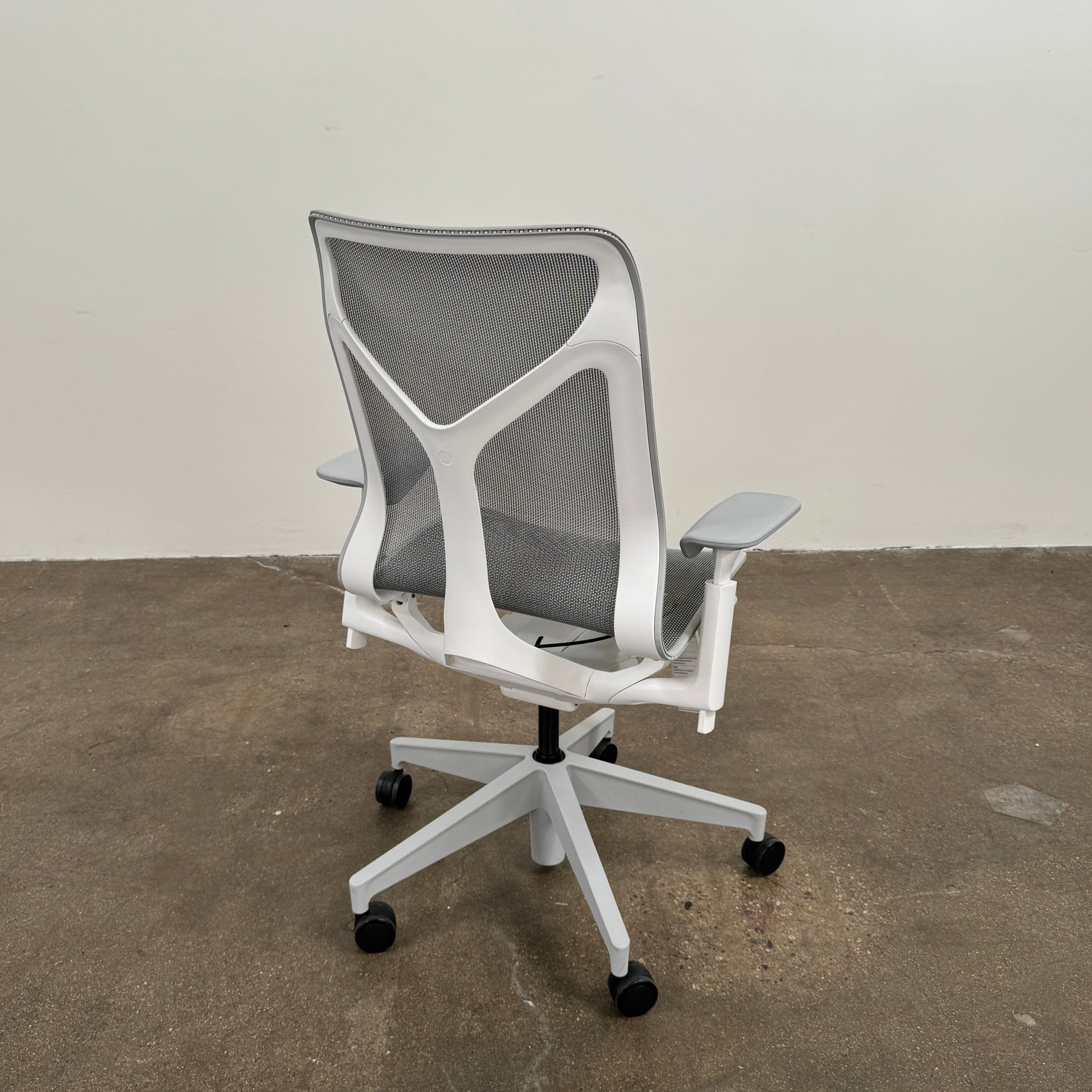 A sleek, modern white Herman Miller Cosm Office Chair featuring a mesh back and seat, supportive armrests, and five caster wheels is showcased on a concrete floor against an off-white wall. Designed by Herman Miller, this ergonomic chair seamlessly combines contemporary style with exceptional comfort.