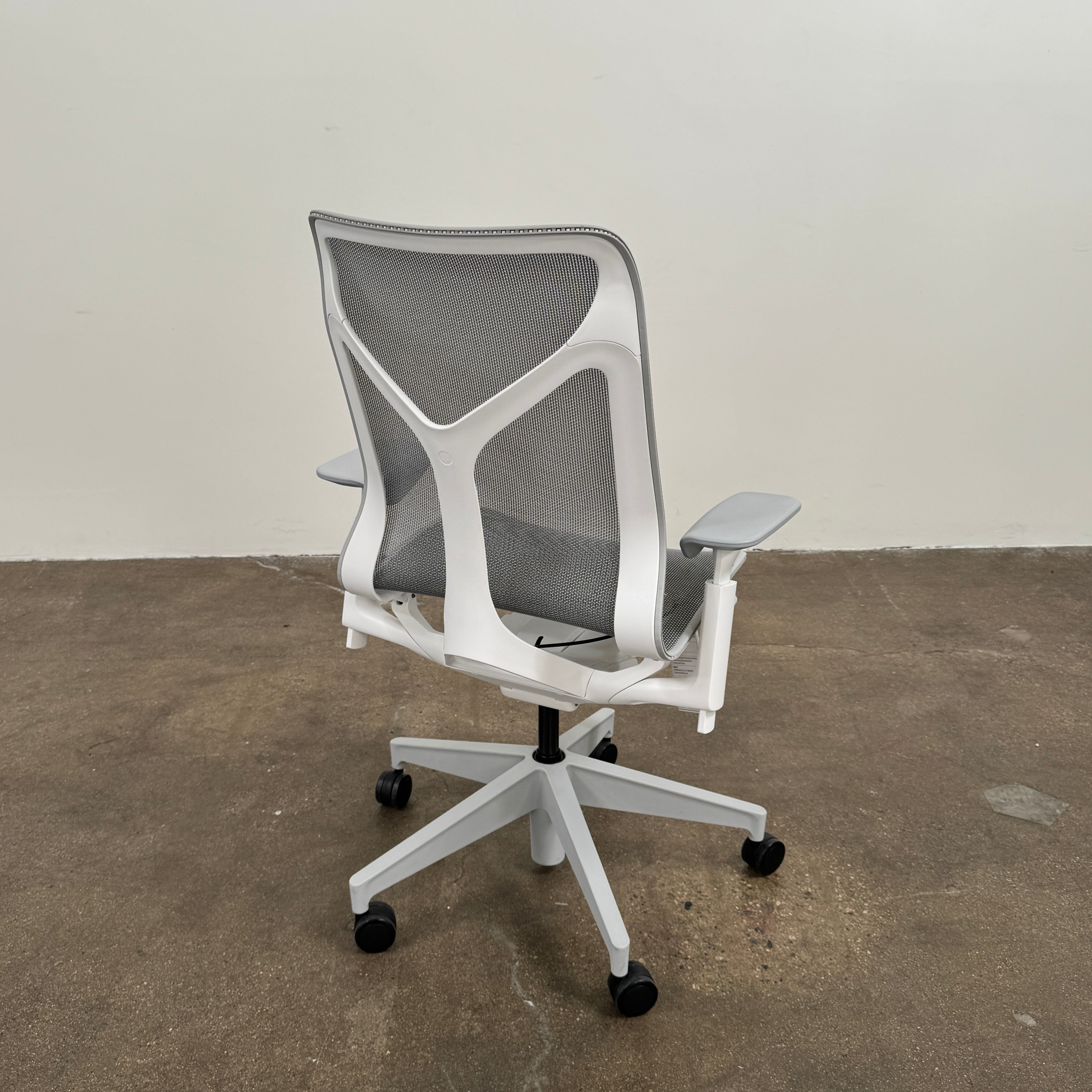 A sleek, modern white Herman Miller Cosm Office Chair featuring a mesh back and seat, supportive armrests, and five caster wheels is showcased on a concrete floor against an off-white wall. Designed by Herman Miller, this ergonomic chair seamlessly combines contemporary style with exceptional comfort.
