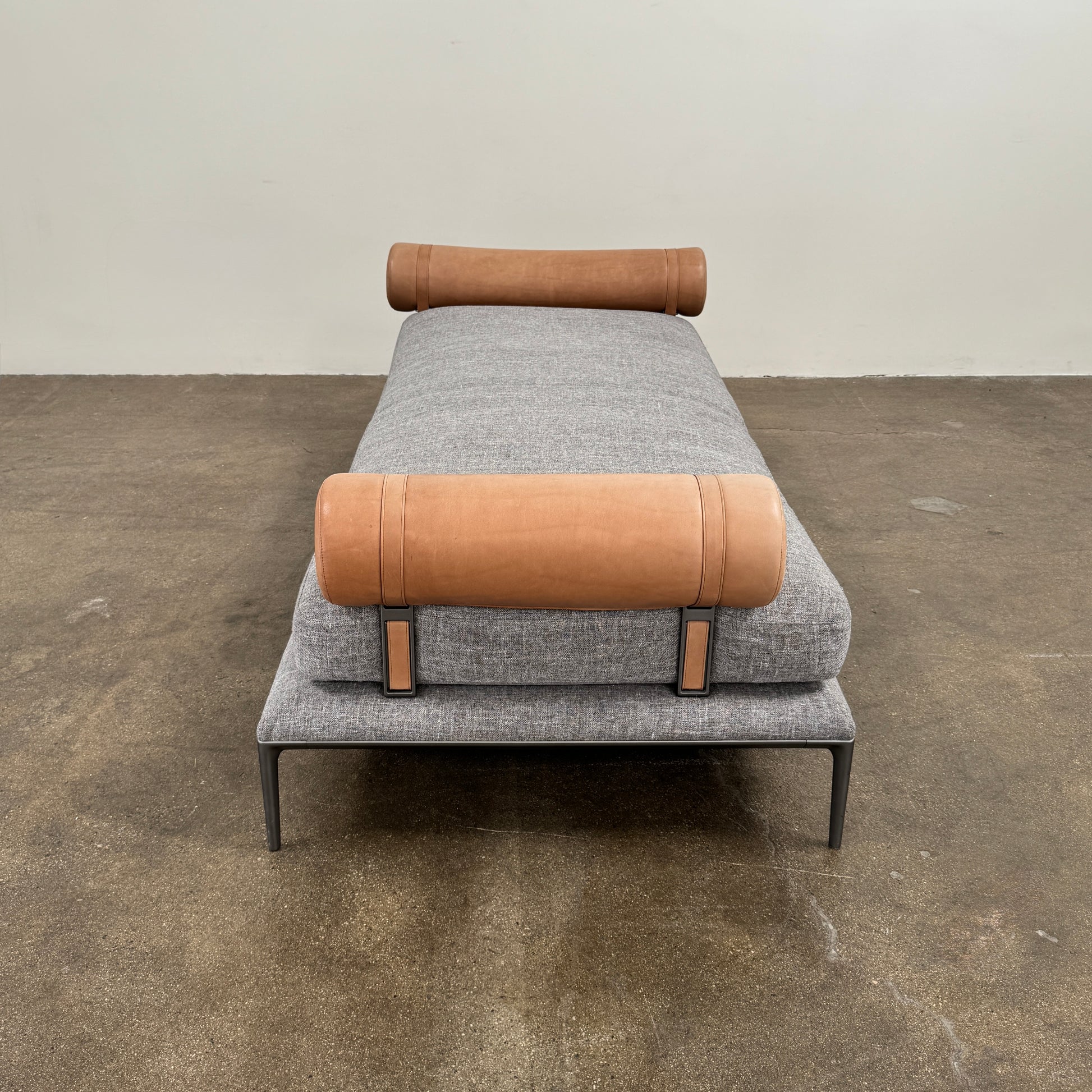 The B&B Italia Atoll Bench with Leather Bolsters by B&B Italia is a modern gray upholstered daybed featuring two cylindrical tan leather bolsters at each end, set on a sleek black metal frame and shown against a white background.