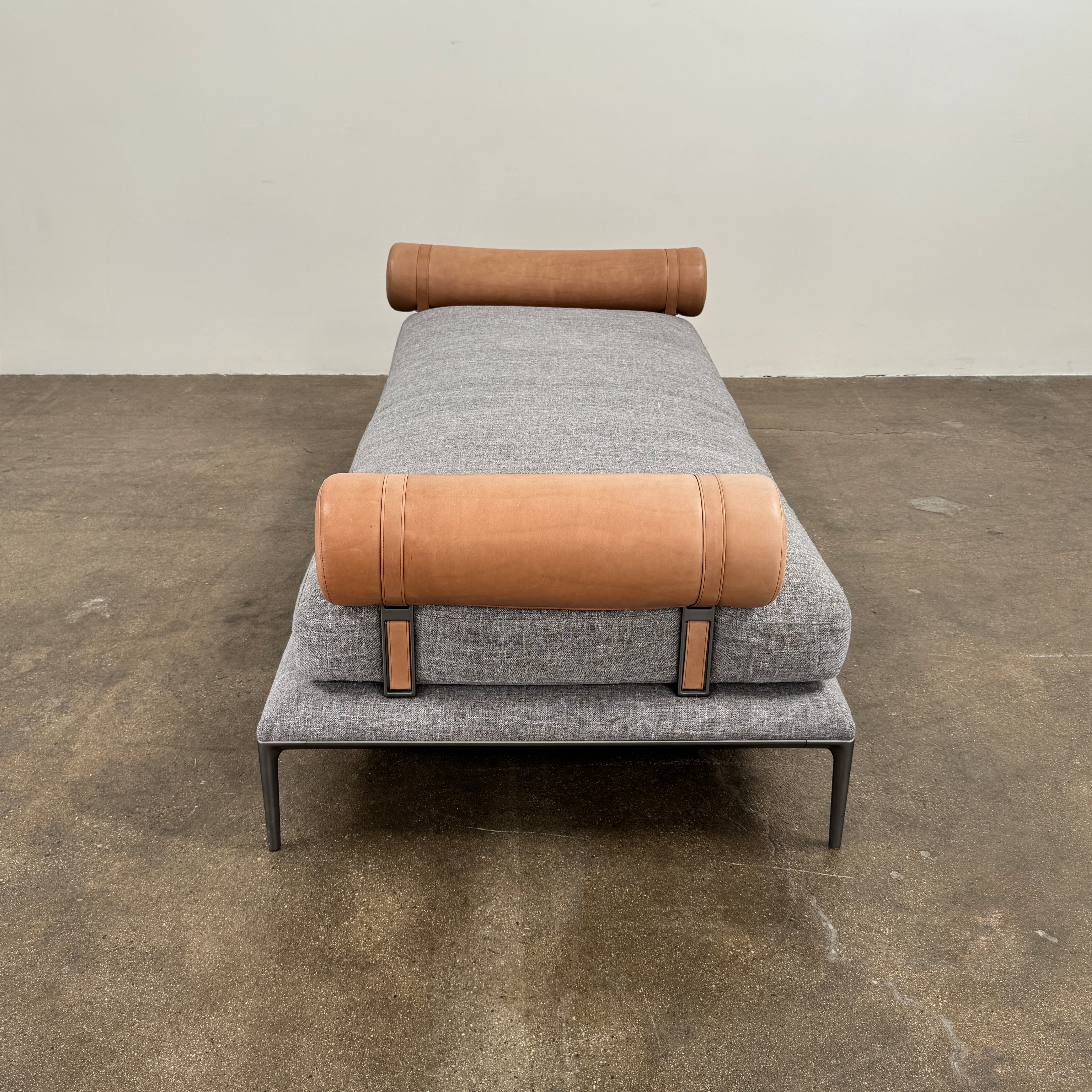 The B&B Italia Atoll Bench with Leather Bolsters by B&B Italia is a modern gray upholstered daybed featuring two cylindrical tan leather bolsters at each end, set on a sleek black metal frame and shown against a white background.