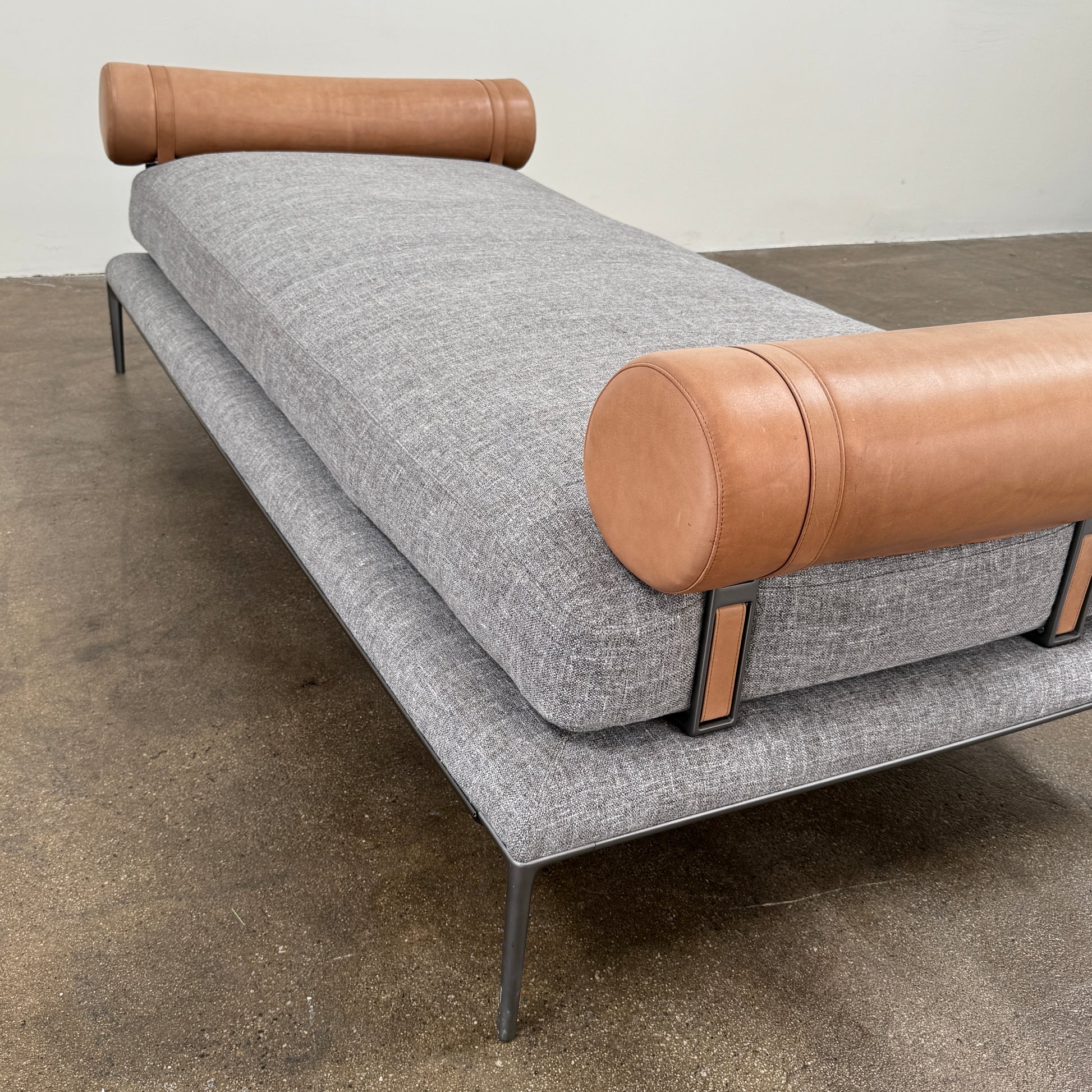 The B&B Italia Atoll Bench with Leather Bolsters by B&B Italia is a modern gray upholstered daybed featuring two cylindrical tan leather bolsters at each end, set on a sleek black metal frame and shown against a white background.