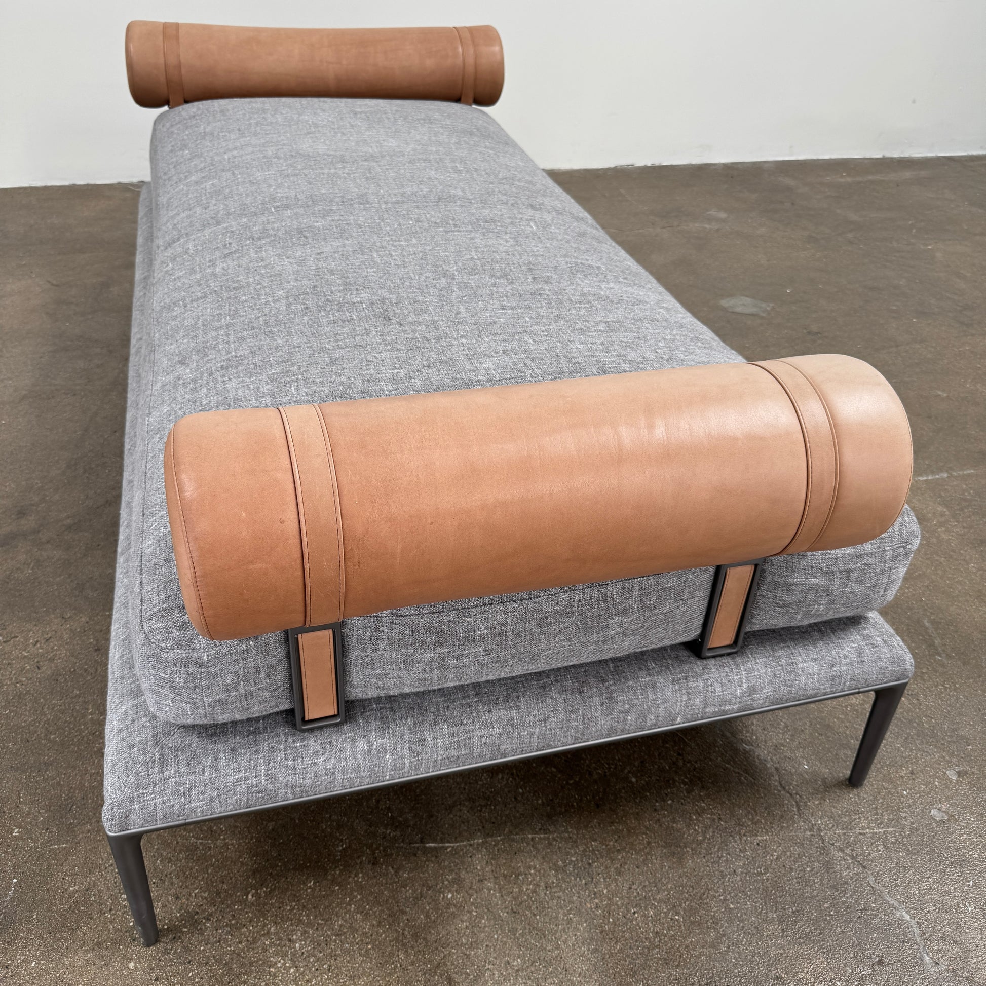 The B&B Italia Atoll Bench with Leather Bolsters by B&B Italia is a modern gray upholstered daybed featuring two cylindrical tan leather bolsters at each end, set on a sleek black metal frame and shown against a white background.