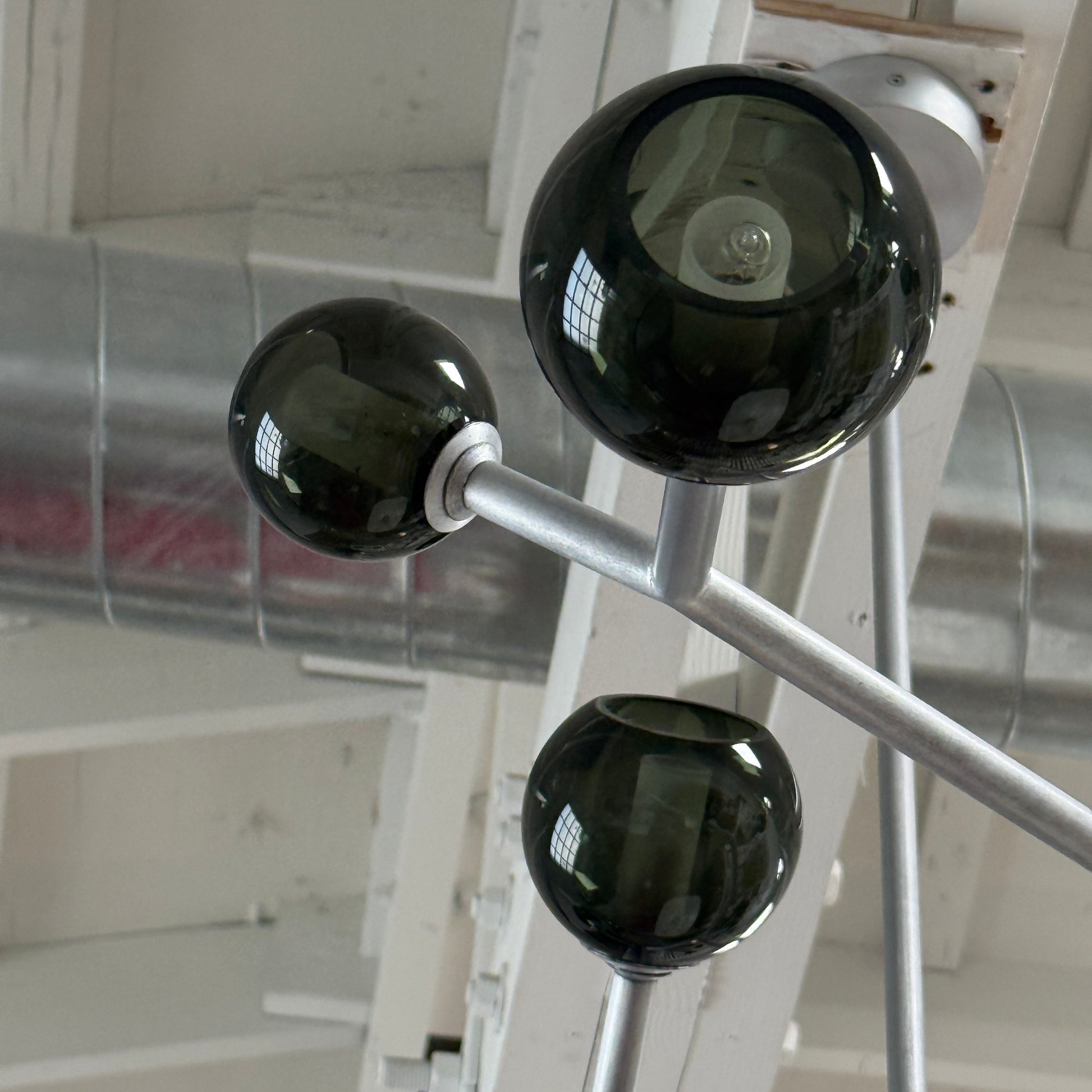 The Pouenat Last Night Branche Chandelier by Pouenat, featuring mouth-blown glass spheres, hangs beneath a white exposed beam ceiling with visible ductwork—an ideal statement for modern industrial interiors.
