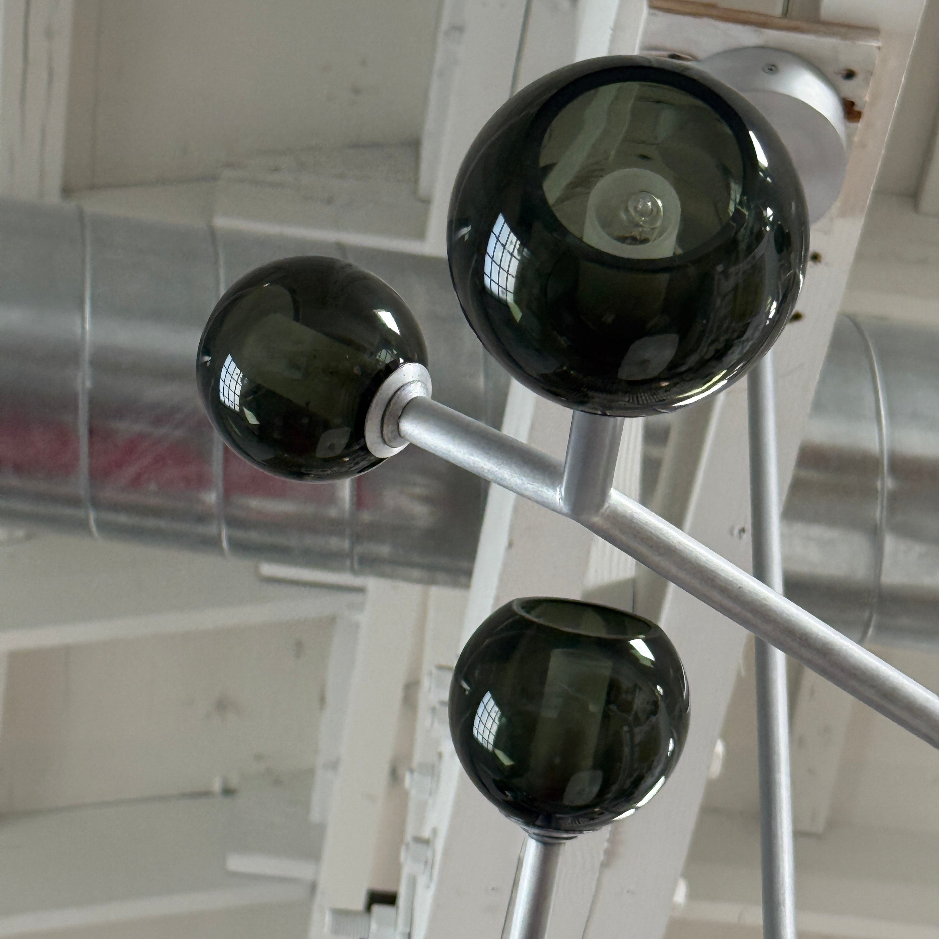 The Pouenat Last Night Branche Chandelier by Pouenat, featuring mouth-blown glass spheres, hangs beneath a white exposed beam ceiling with visible ductwork—an ideal statement for modern industrial interiors.