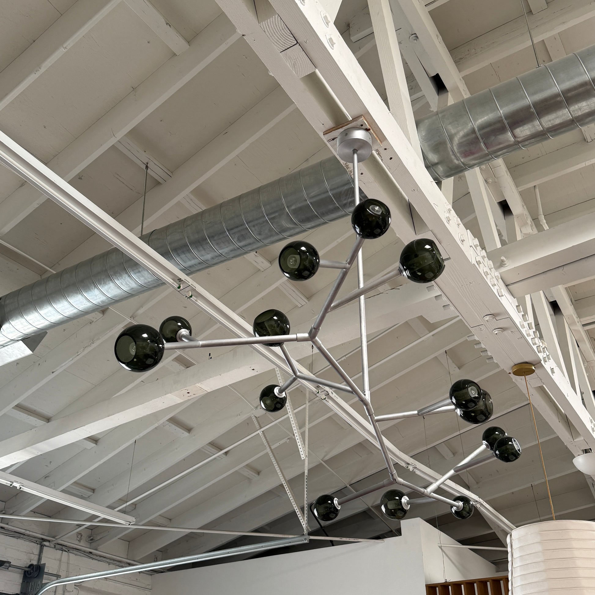 The Pouenat Last Night Branche Chandelier by Pouenat, featuring mouth-blown glass spheres, hangs beneath a white exposed beam ceiling with visible ductwork—an ideal statement for modern industrial interiors.
