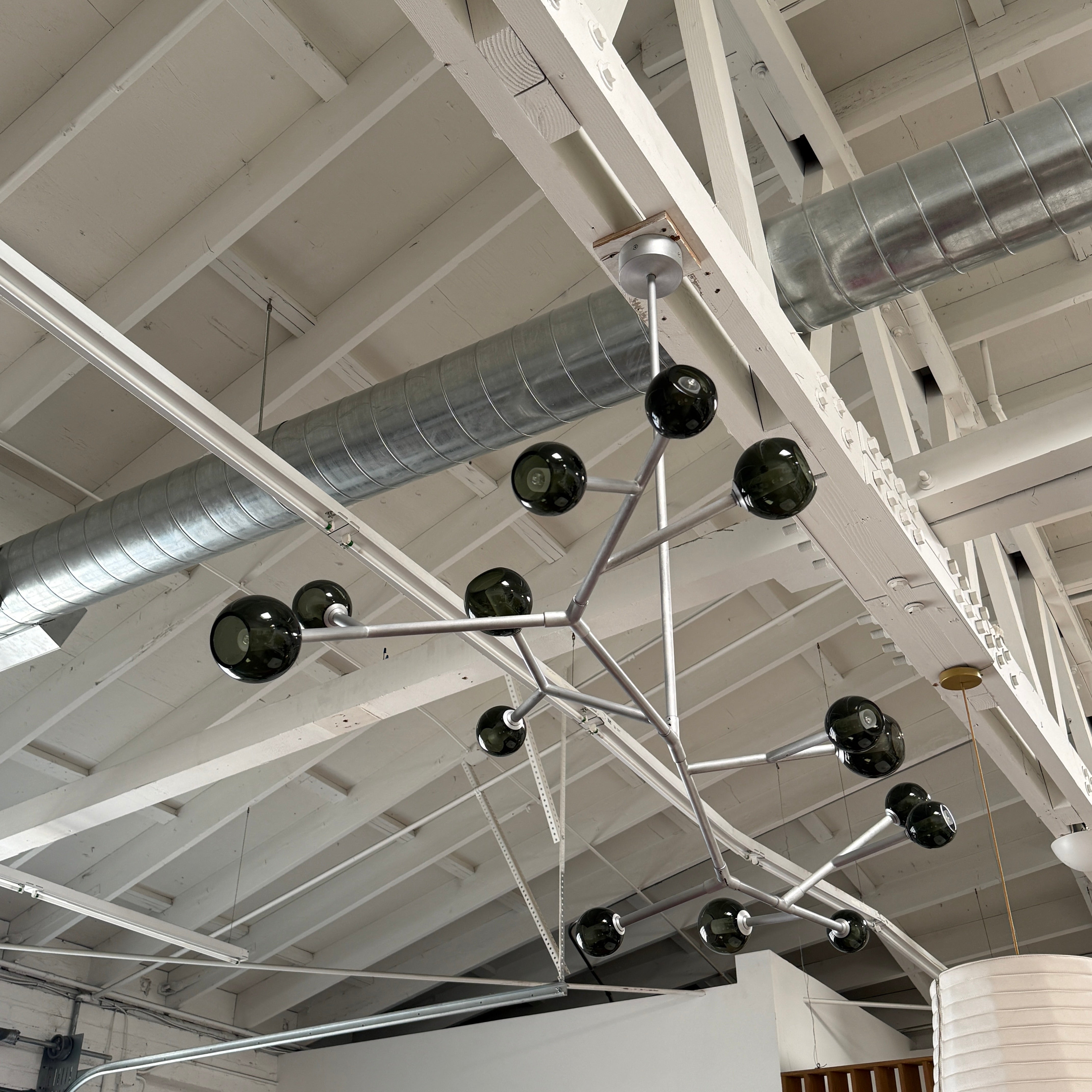 The Pouenat Last Night Branche Chandelier by Pouenat, featuring mouth-blown glass spheres, hangs beneath a white exposed beam ceiling with visible ductwork—an ideal statement for modern industrial interiors.