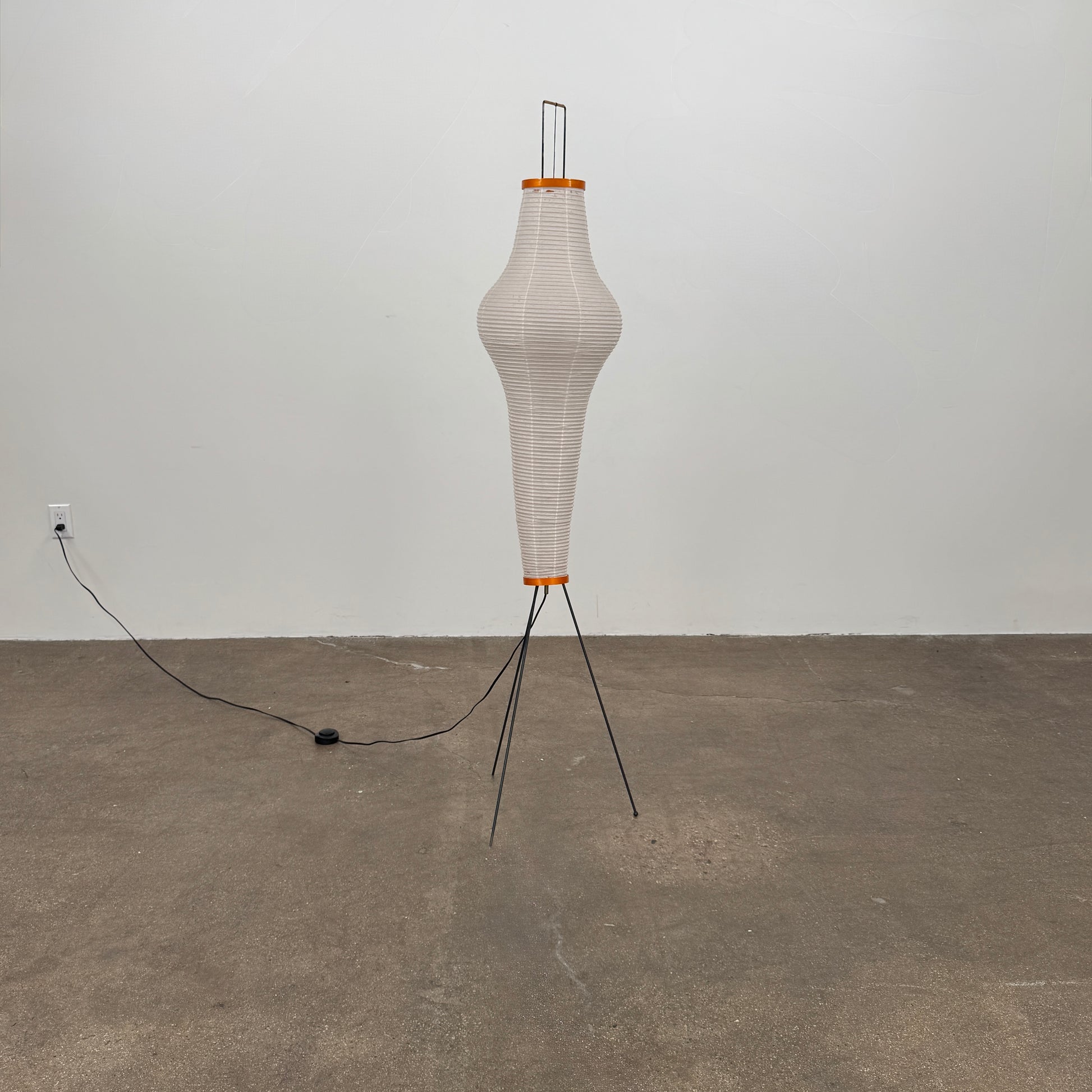 The Noguchi Araki 14A Floor Lamp by Noguchi is a tall, sculptural floor lamp inspired by Japanese craftsmanship, featuring a white ribbed shade, wooden accents, and three slender metal legs. Reminiscent of Akari Light Sculptures and the original Noguchi Araki 14A design, this plugged-in lamp is shown illuminated against a plain white background.