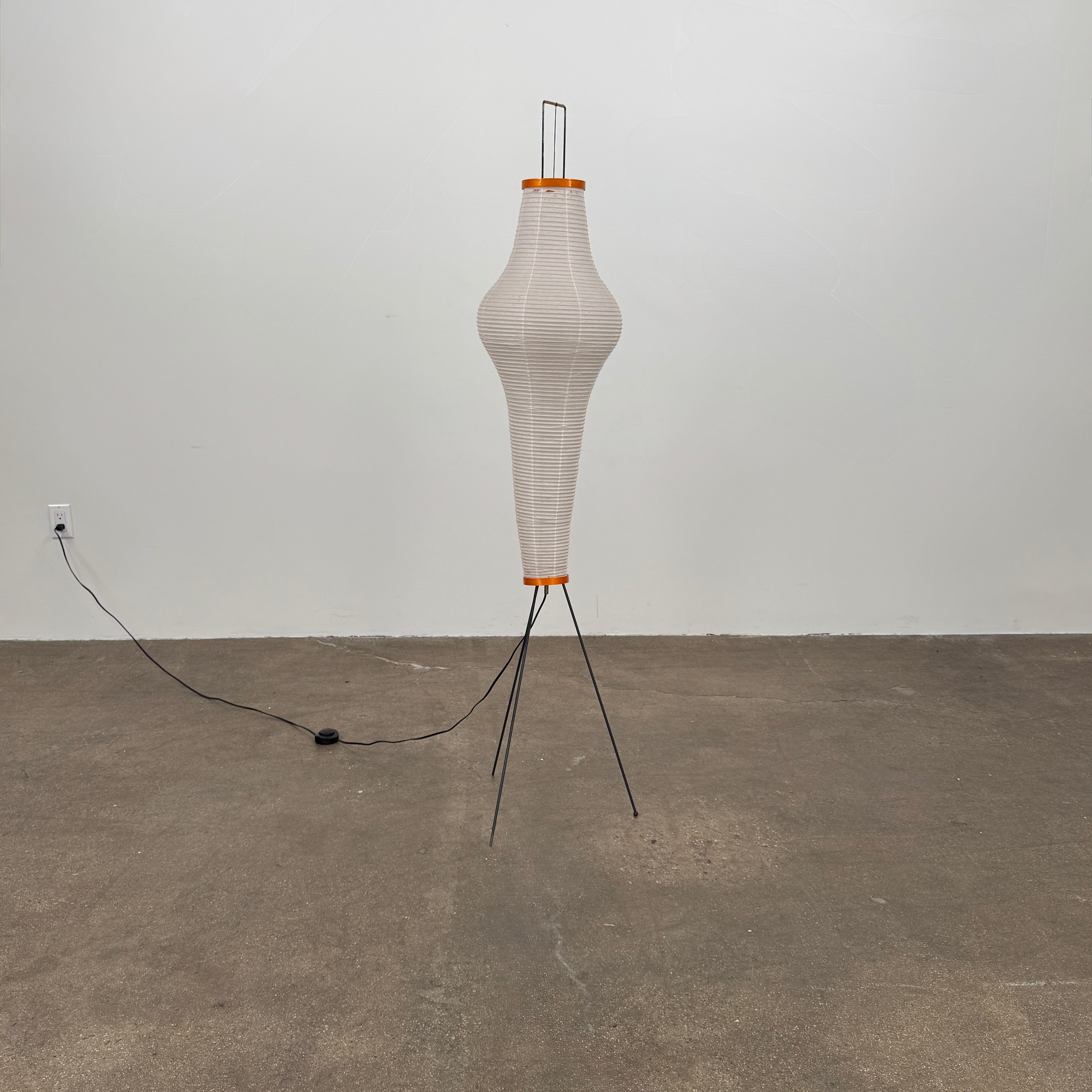 The Noguchi Araki 14A Floor Lamp by Noguchi is a tall, sculptural floor lamp inspired by Japanese craftsmanship, featuring a white ribbed shade, wooden accents, and three slender metal legs. Reminiscent of Akari Light Sculptures and the original Noguchi Araki 14A design, this plugged-in lamp is shown illuminated against a plain white background.