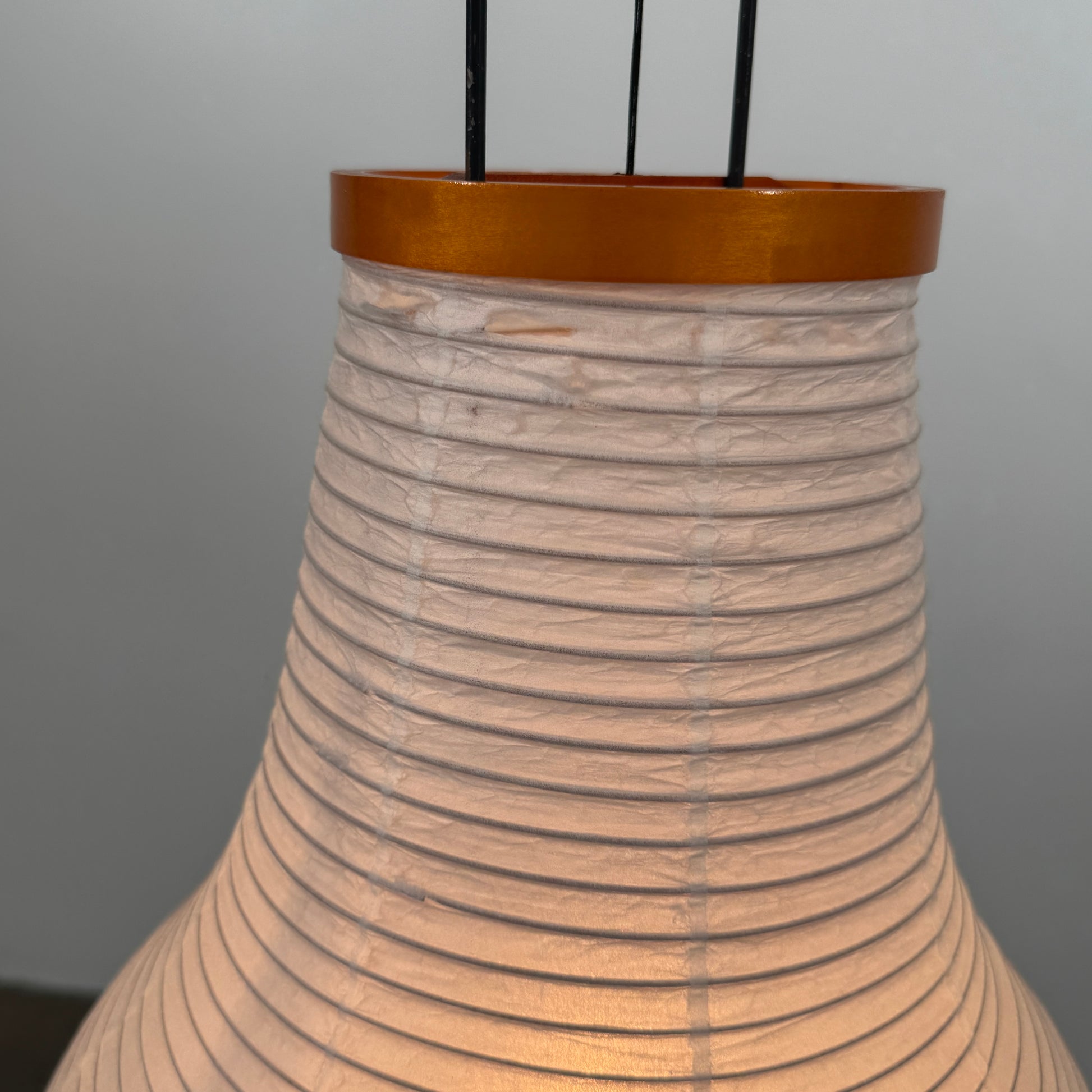 The Noguchi Araki 14A Floor Lamp by Noguchi is a tall, sculptural floor lamp inspired by Japanese craftsmanship, featuring a white ribbed shade, wooden accents, and three slender metal legs. Reminiscent of Akari Light Sculptures and the original Noguchi Araki 14A design, this plugged-in lamp is shown illuminated against a plain white background.