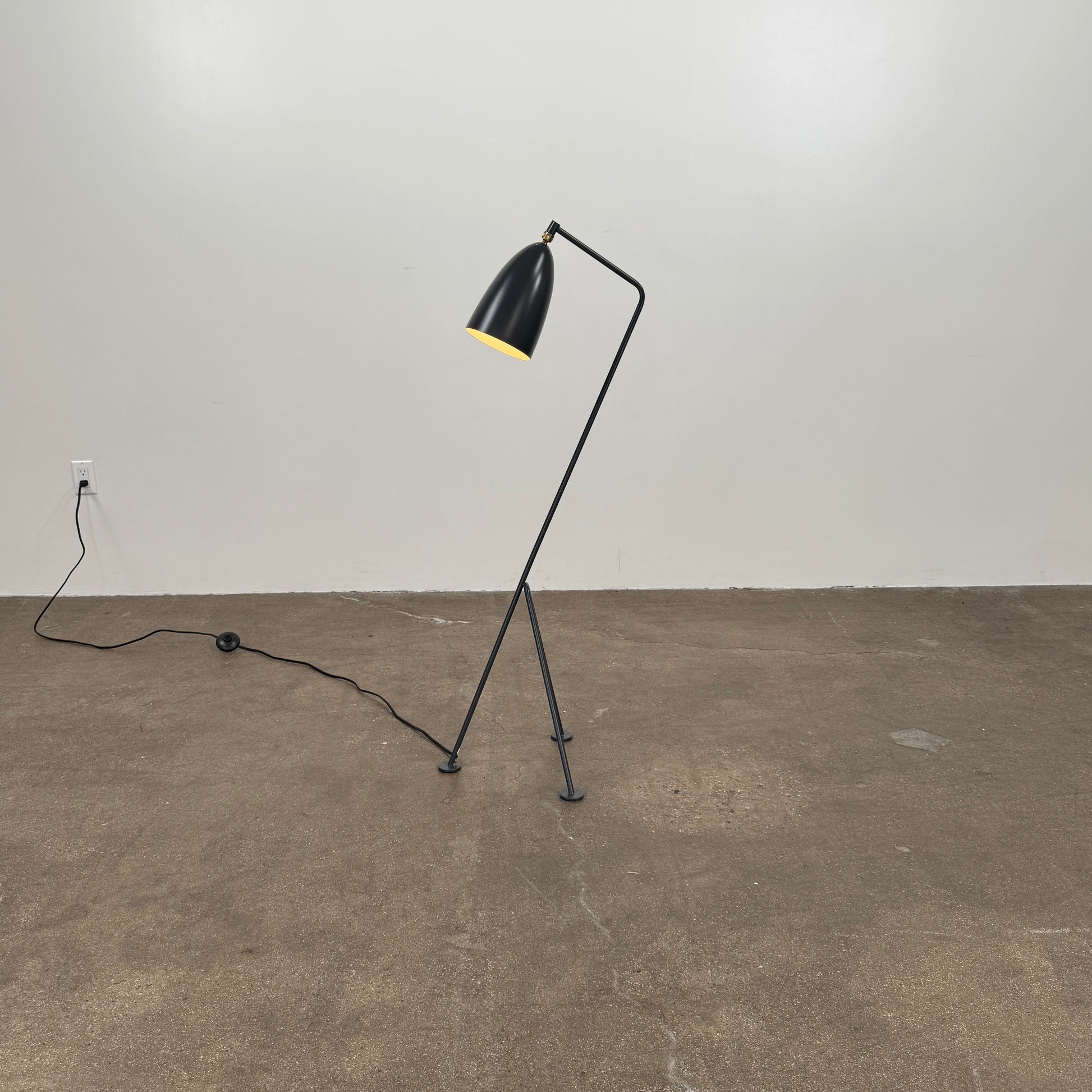 The Gubi Grashoppa Floor Lamp by Gubi, featuring a black finish, curved neck, tripod base, and bell-shaped shade inspired by the iconic Greta Grossman design, stands on a white background with its power cord visible.