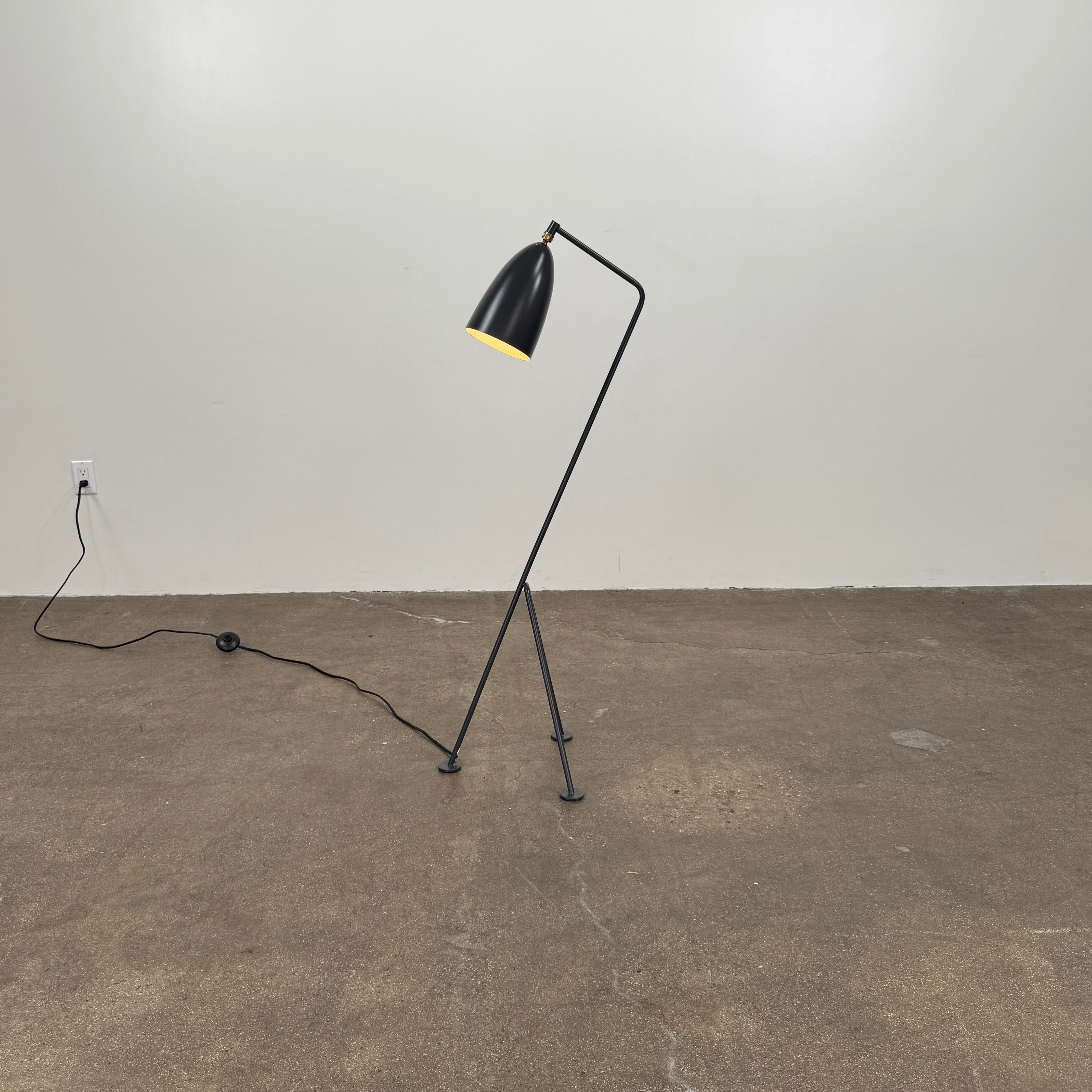 The Gubi Grashoppa Floor Lamp by Gubi, featuring a black finish, curved neck, tripod base, and bell-shaped shade inspired by the iconic Greta Grossman design, stands on a white background with its power cord visible.
