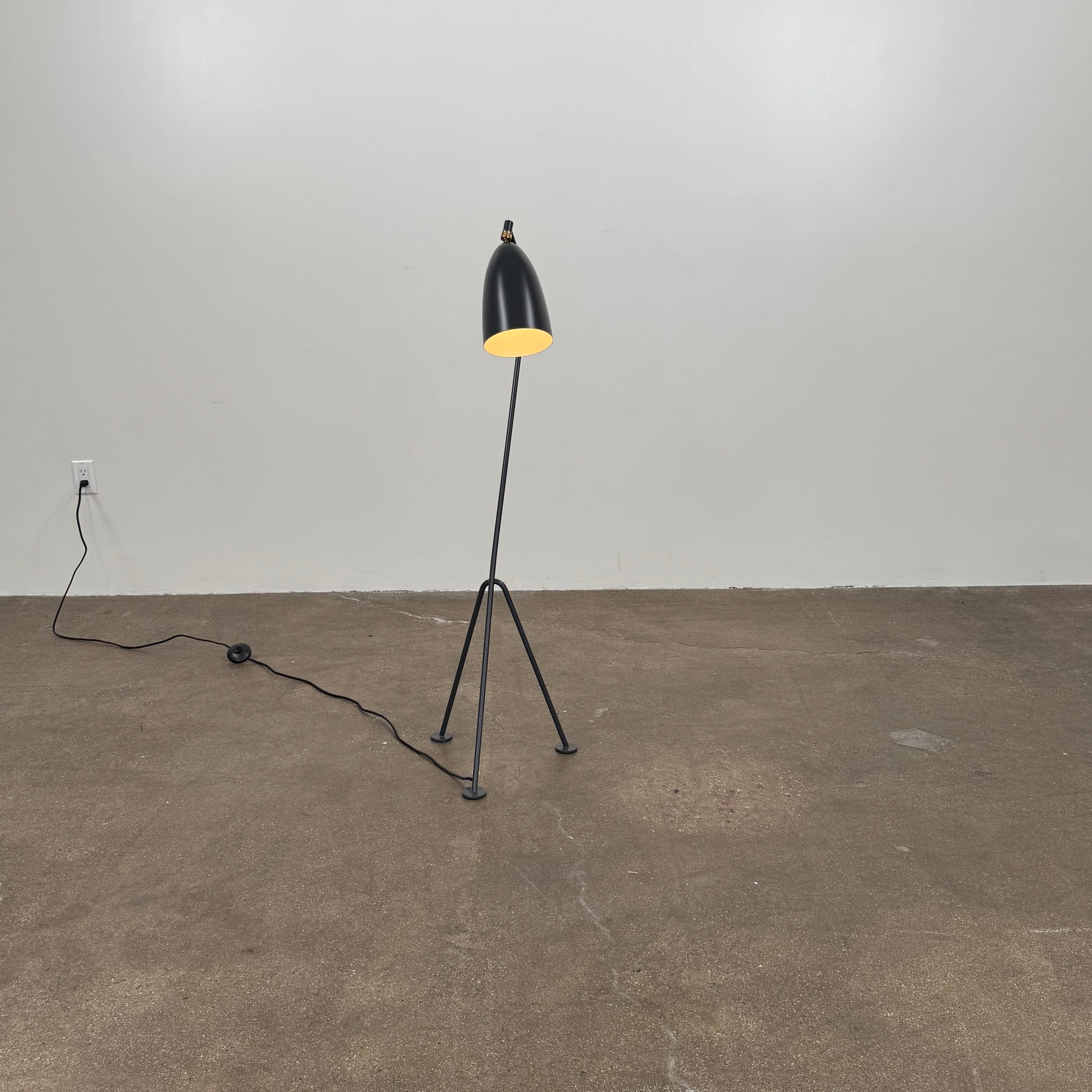 The Gubi Grashoppa Floor Lamp by Gubi, featuring a black finish, curved neck, tripod base, and bell-shaped shade inspired by the iconic Greta Grossman design, stands on a white background with its power cord visible.