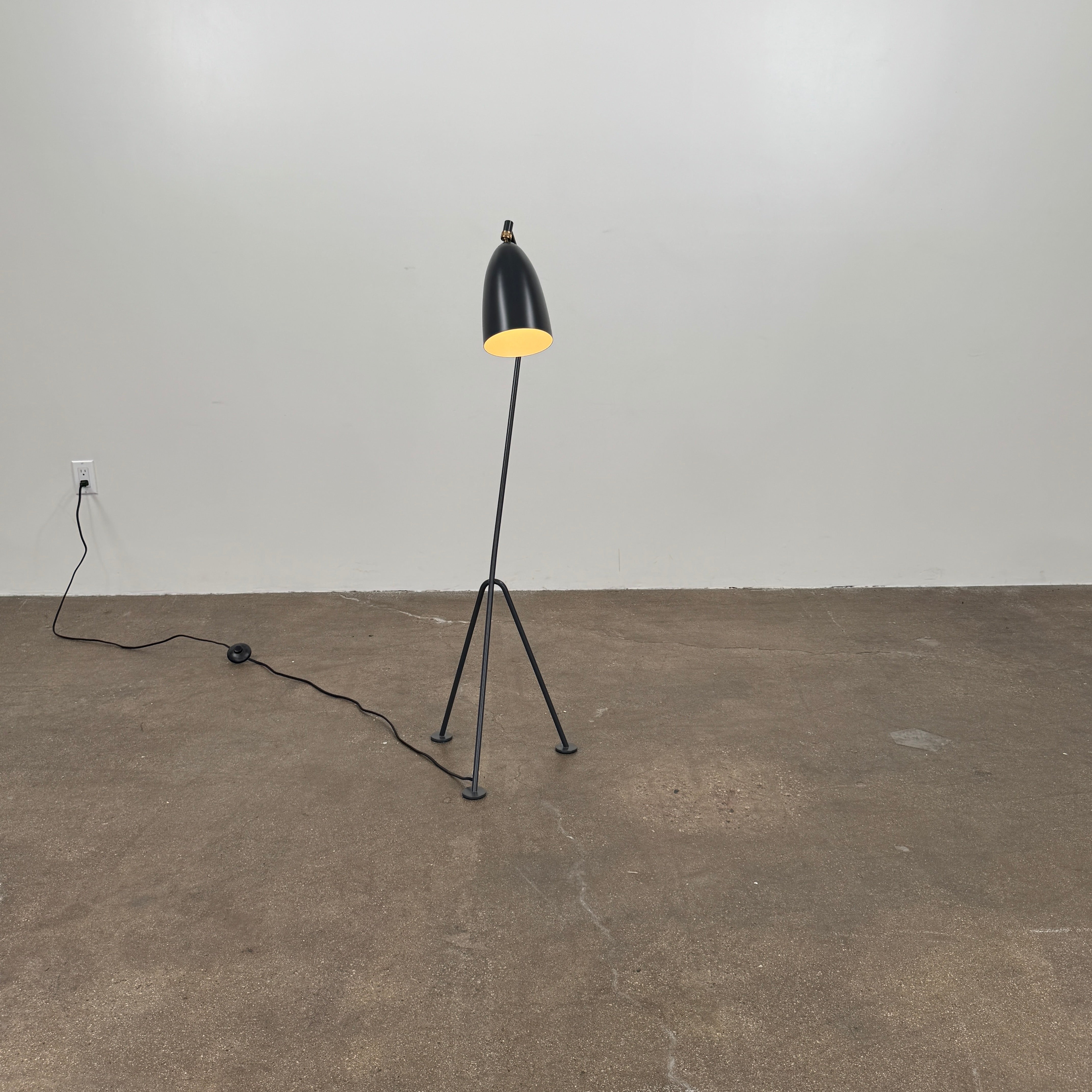 The Gubi Grashoppa Floor Lamp by Gubi, featuring a black finish, curved neck, tripod base, and bell-shaped shade inspired by the iconic Greta Grossman design, stands on a white background with its power cord visible.