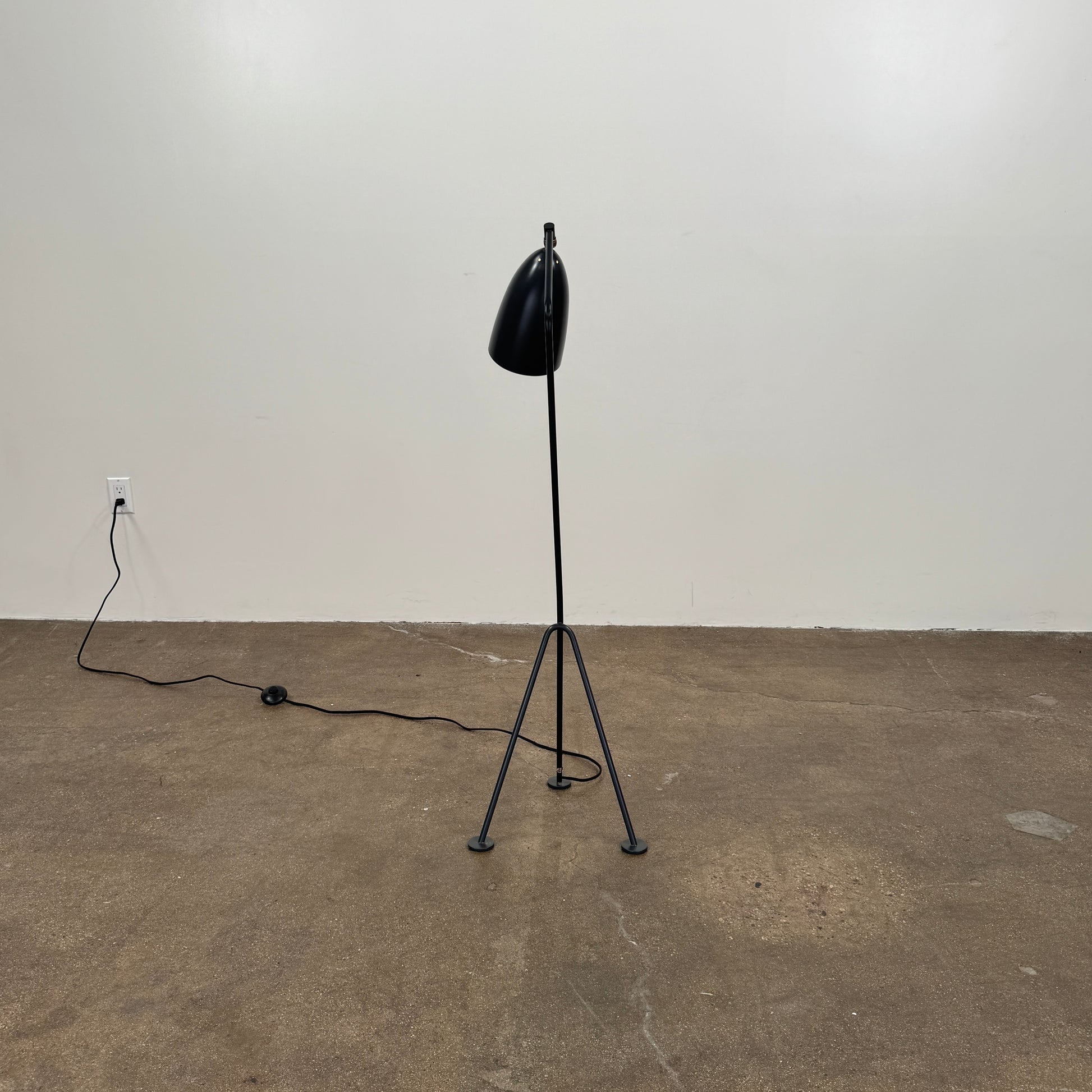 The Gubi Grashoppa Floor Lamp by Gubi, featuring a black finish, curved neck, tripod base, and bell-shaped shade inspired by the iconic Greta Grossman design, stands on a white background with its power cord visible.