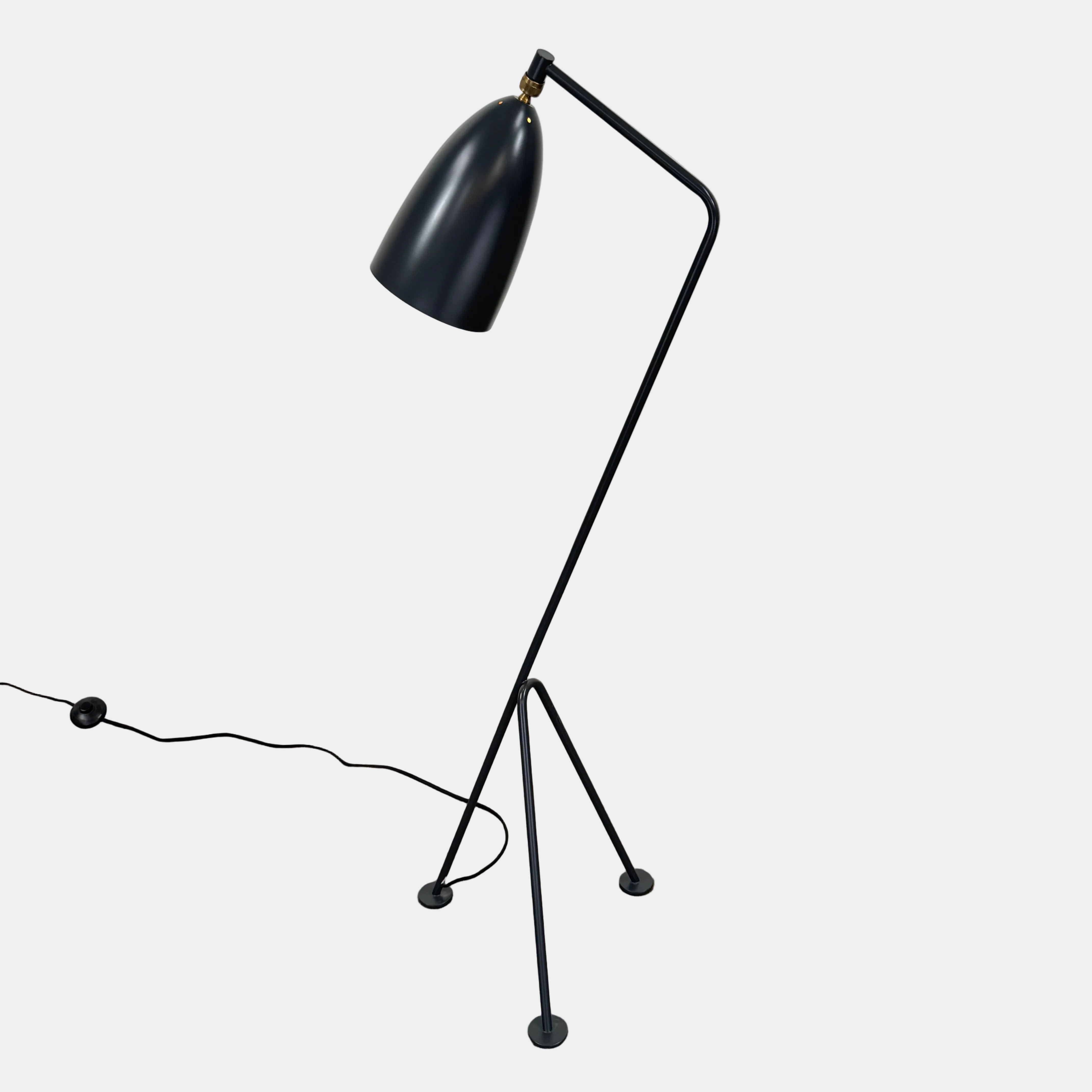 The Gubi Grashoppa Floor Lamp by Gubi, featuring a black finish, curved neck, tripod base, and bell-shaped shade inspired by the iconic Greta Grossman design, stands on a white background with its power cord visible.