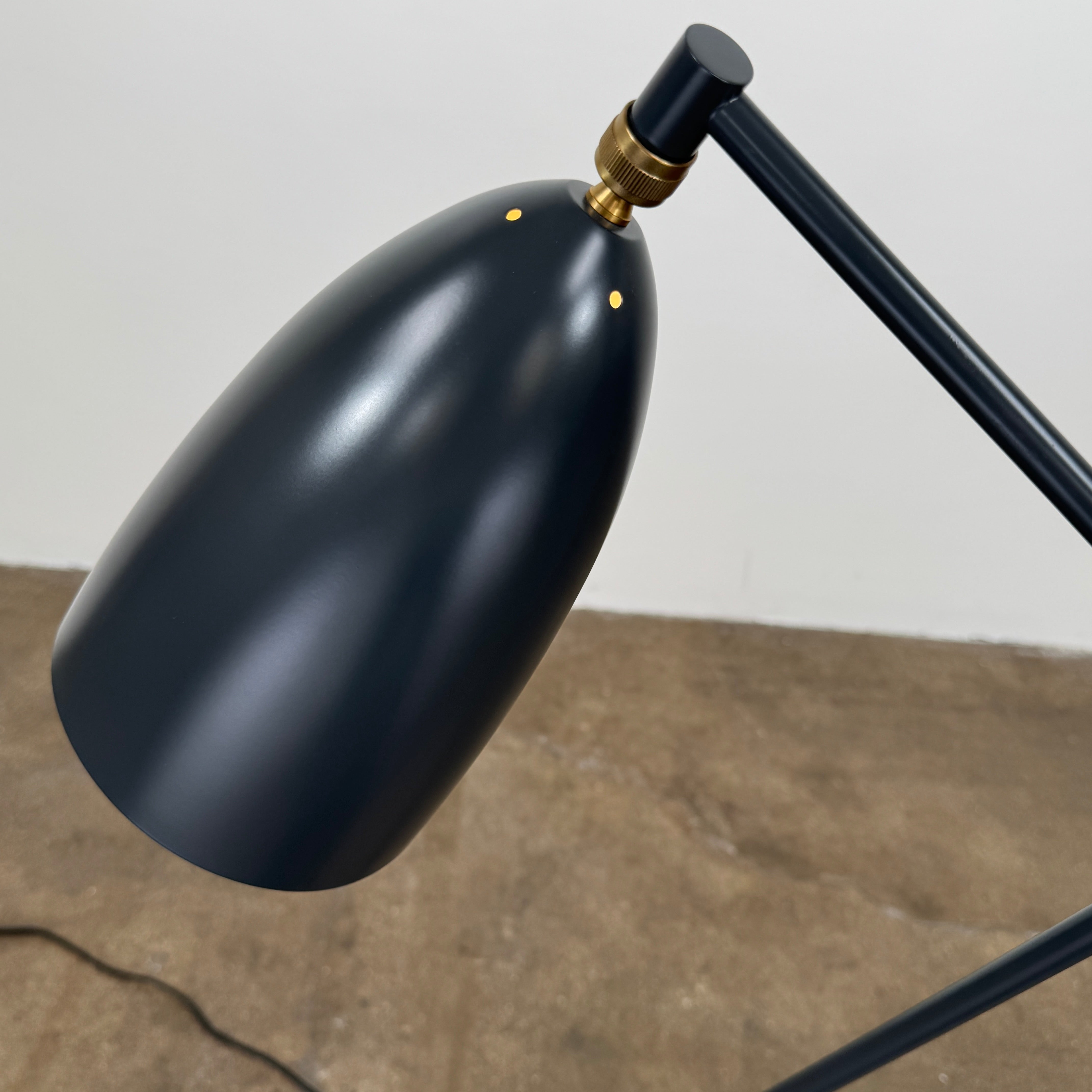 The Gubi Grashoppa Floor Lamp by Gubi, featuring a black finish, curved neck, tripod base, and bell-shaped shade inspired by the iconic Greta Grossman design, stands on a white background with its power cord visible.