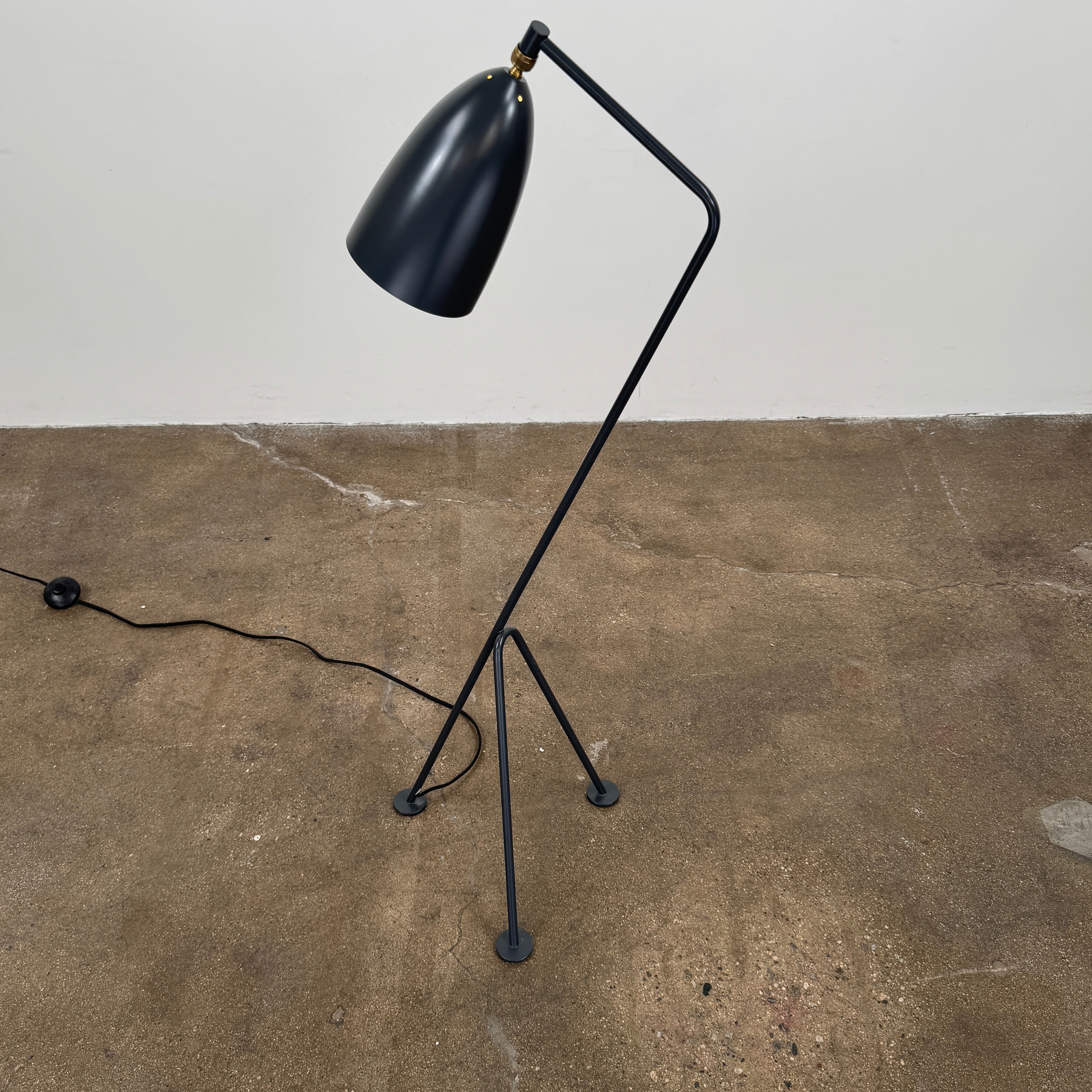 The Gubi Grashoppa Floor Lamp by Gubi, featuring a black finish, curved neck, tripod base, and bell-shaped shade inspired by the iconic Greta Grossman design, stands on a white background with its power cord visible.