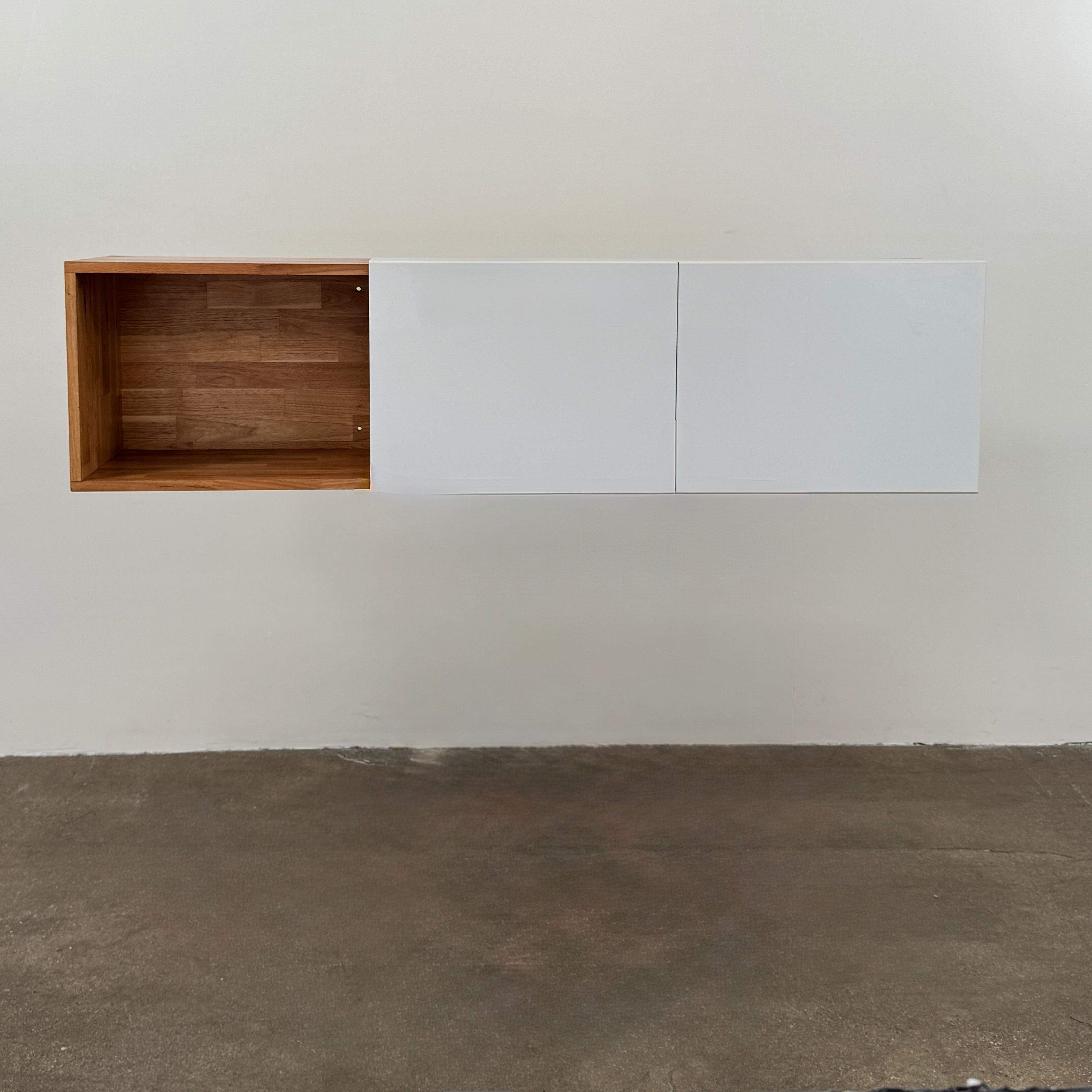 A Modern Resale Mash Studios LAX series 3X Wall Mounted Shelf in natural ash wood and matte white is mounted against a plain wall above a concrete floor, featuring a white exterior with wood-toned interior shelving.